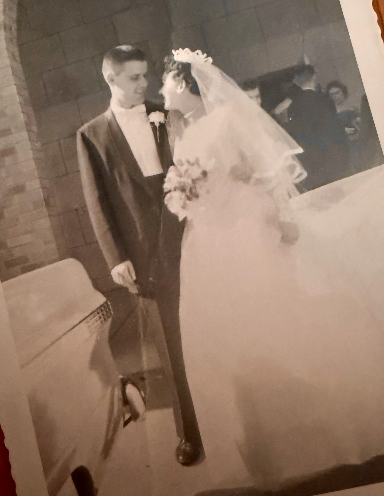 Bride in a long wedding dress and veil stands beside a groom in a suit and tie, holding a bouquet at a wedding ceremony venue.
