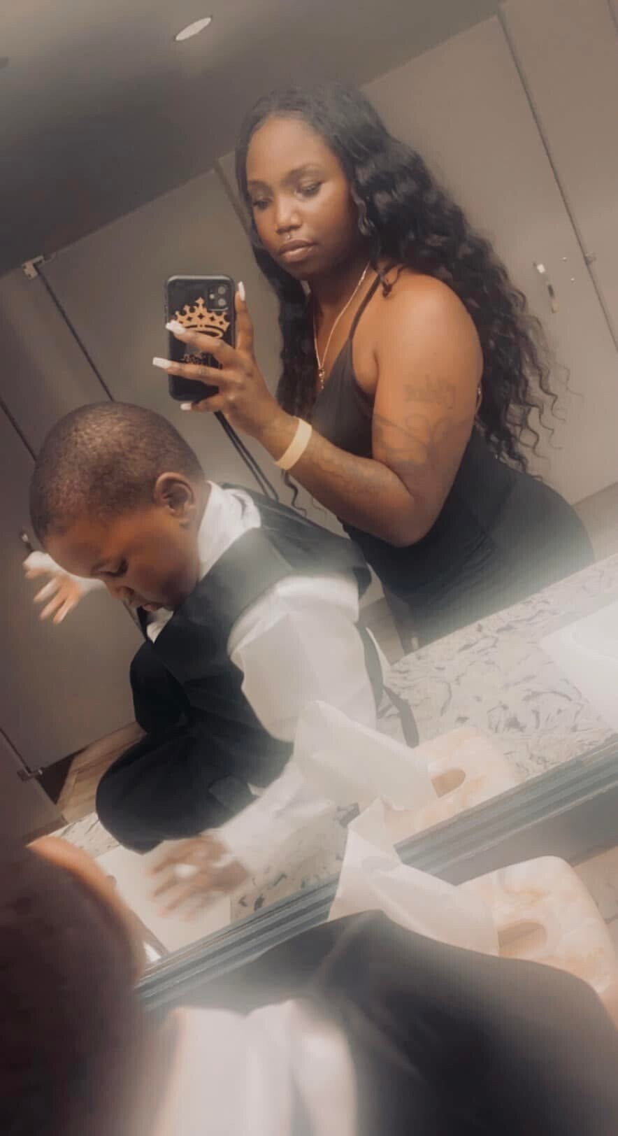Woman with long black hair in a black tank top takes a bathroom mirror selfie; man in a suit sits at the counter.
