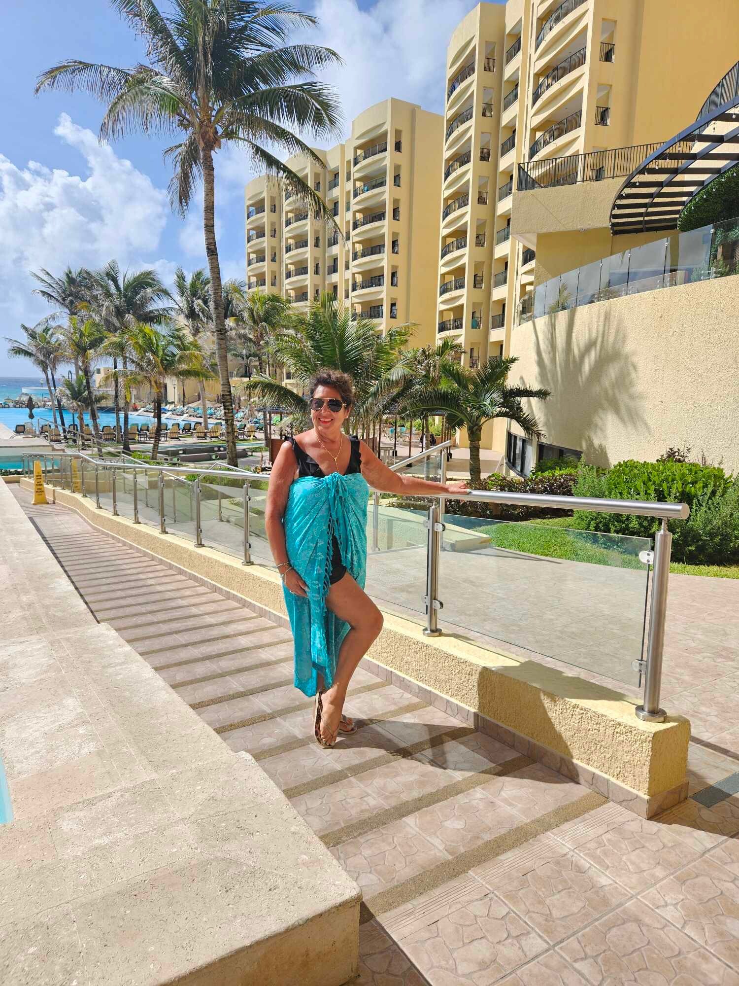 Smiling woman in a turquoise cover-up stands on a resort walkway with palm trees, a pool, and yellow hotel towers in the background.