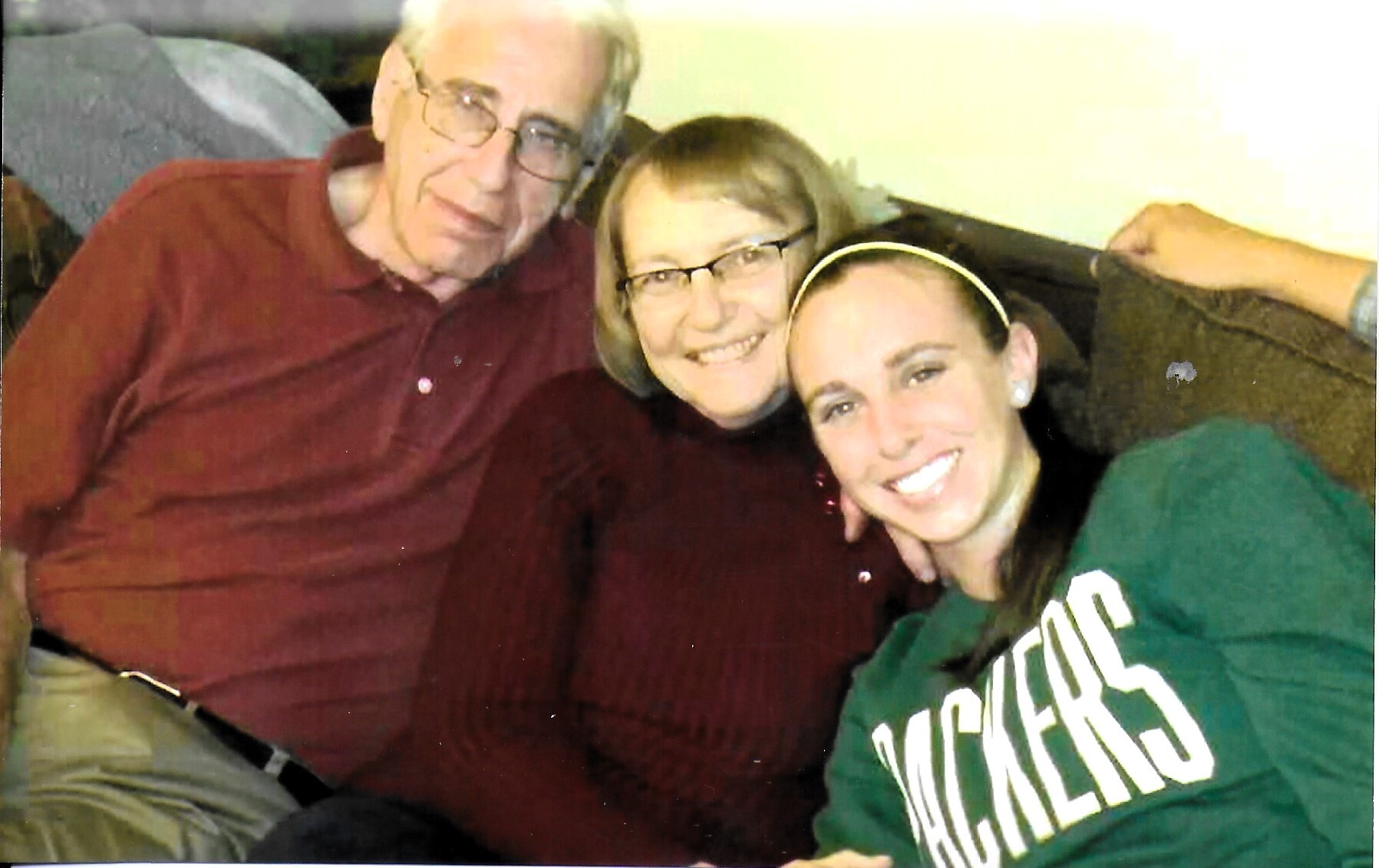 Three people sit closely on a couch, smiling for the camera: an older man in a red shirt, a middle-aged woman, and a young woman in a green Michigan sweatshirt.