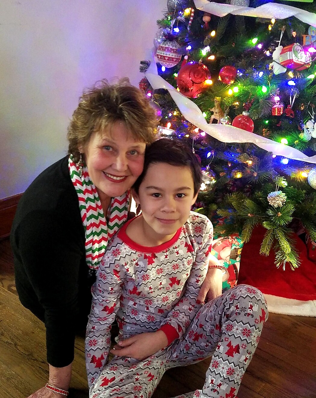 Grandmother and child pose together by a brightly lit Christmas tree with colorful ornaments and ribbons.