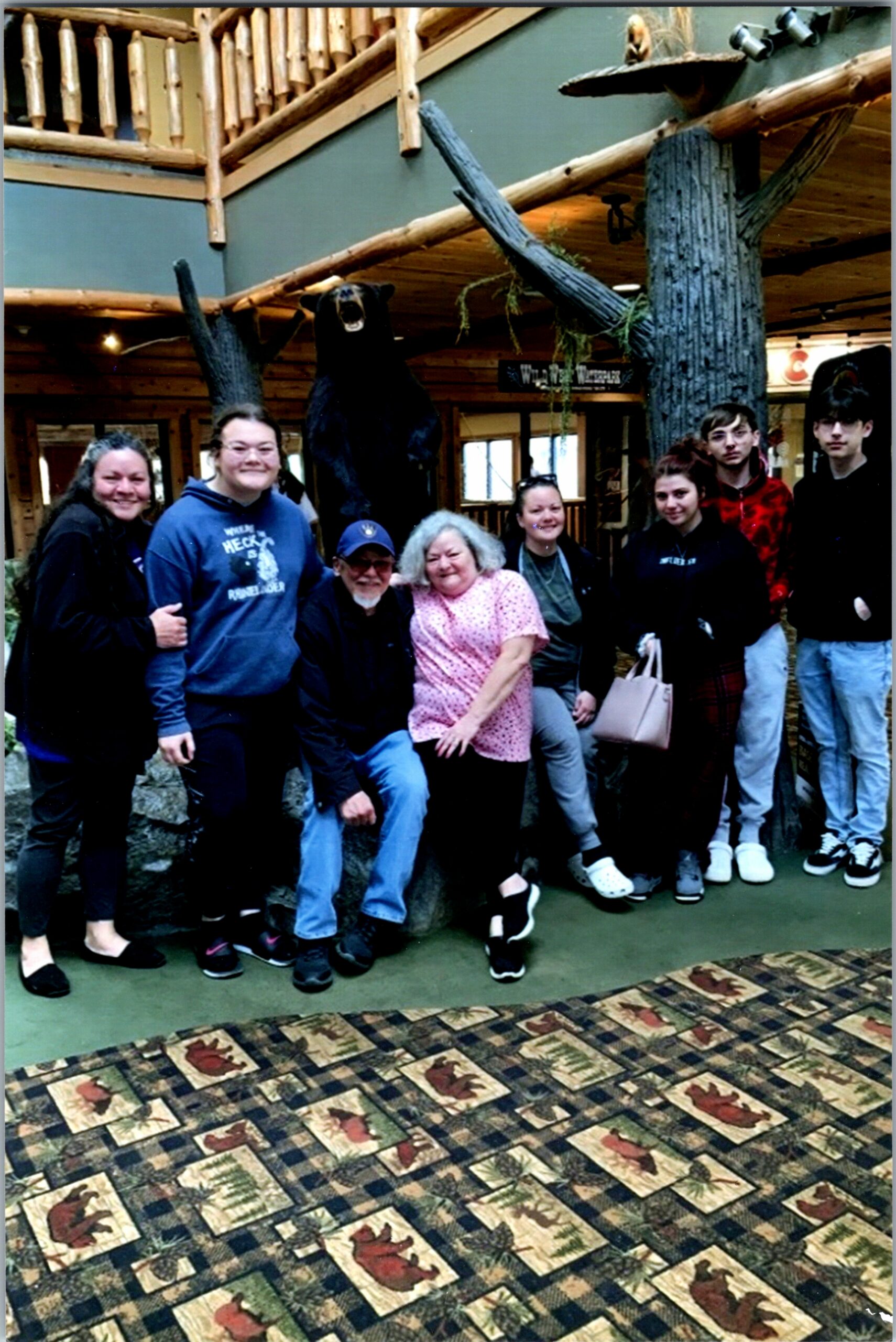 Group of nine people posing and smiling in a rustic lodge, with a large black bear statue in the background and a decorative bear-patterned carpet on the floor.
