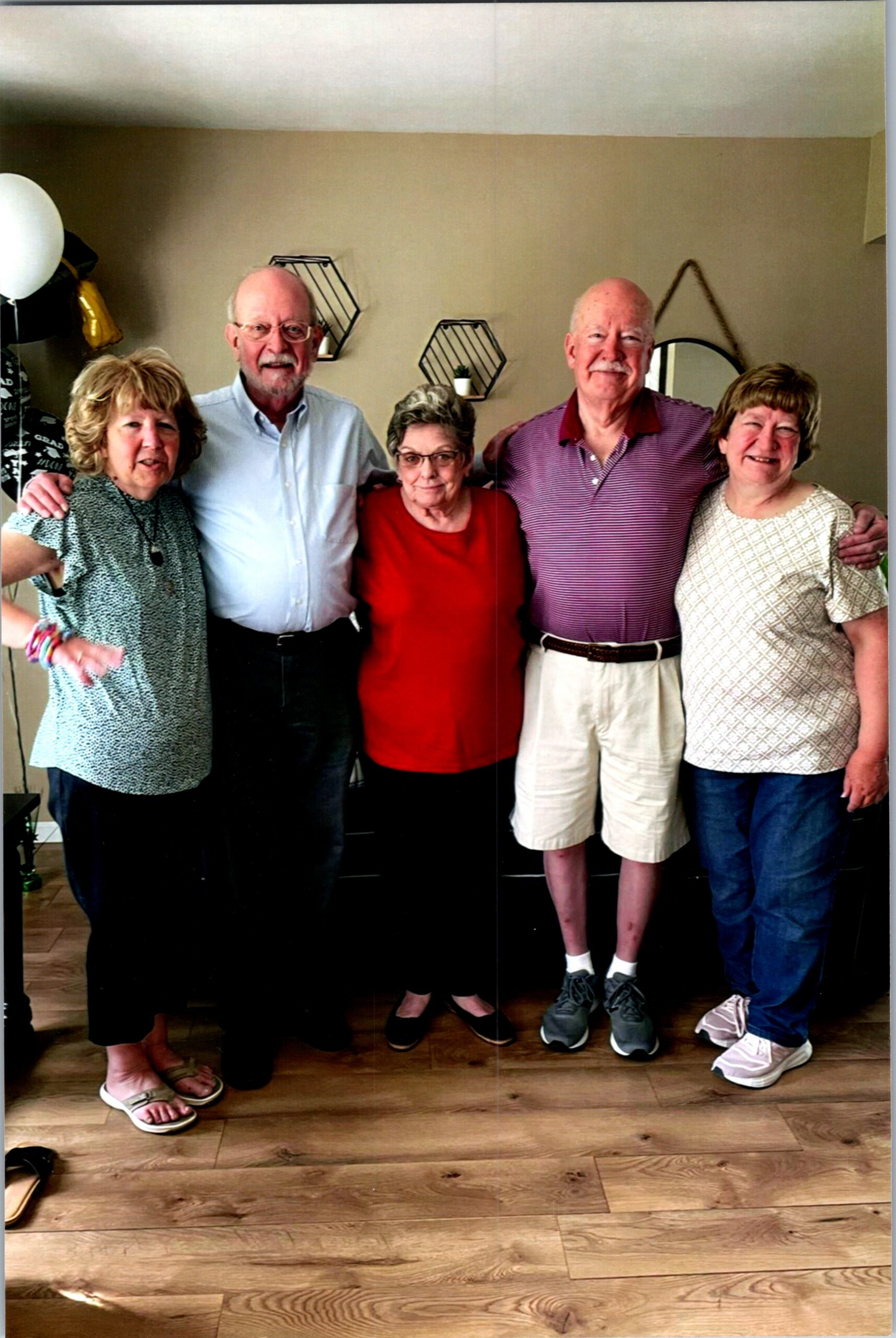 Group of five smiling older adults posing together indoors, arms around each other in a casual home setting with wooden floor and wall decor behind them.