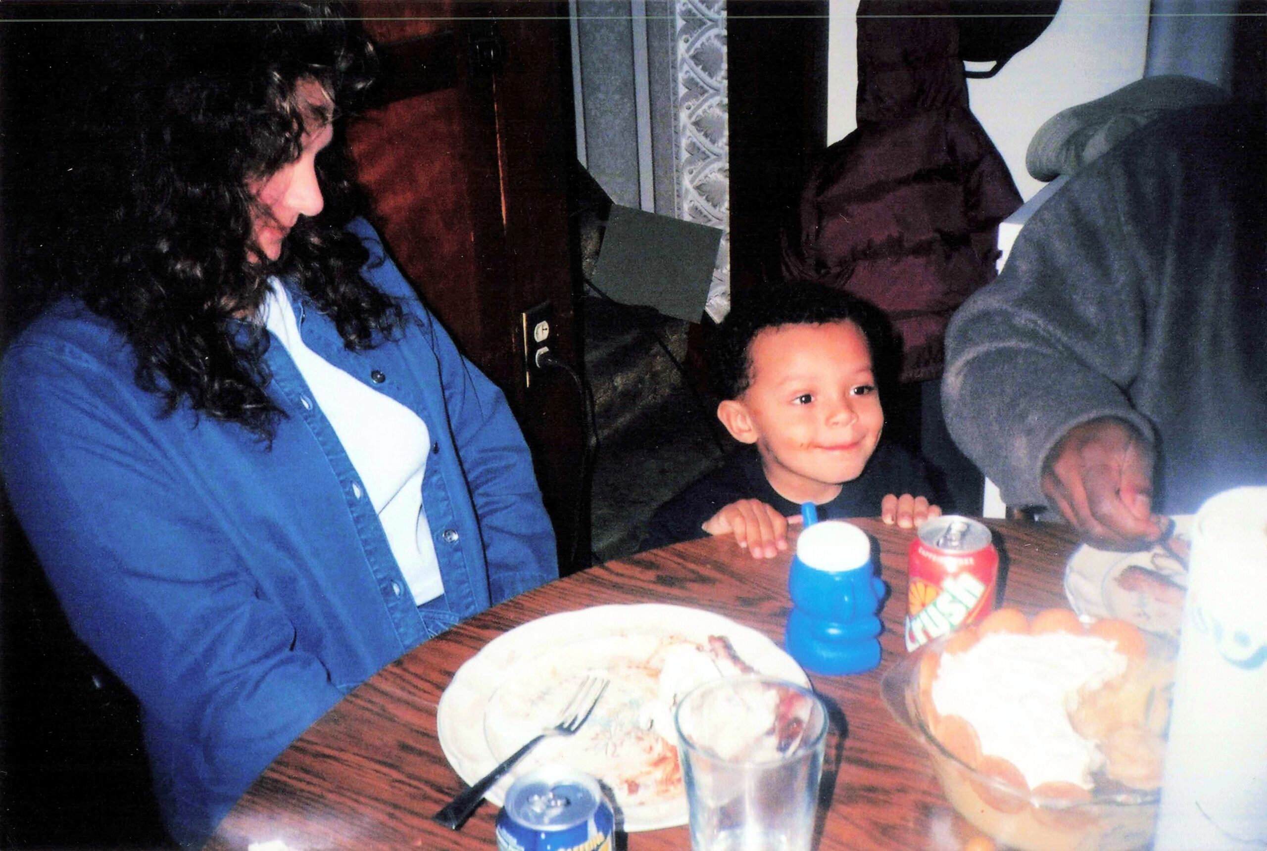 A smiling child sits at a dining table with adults nearby, blue cups and juice boxes in view.