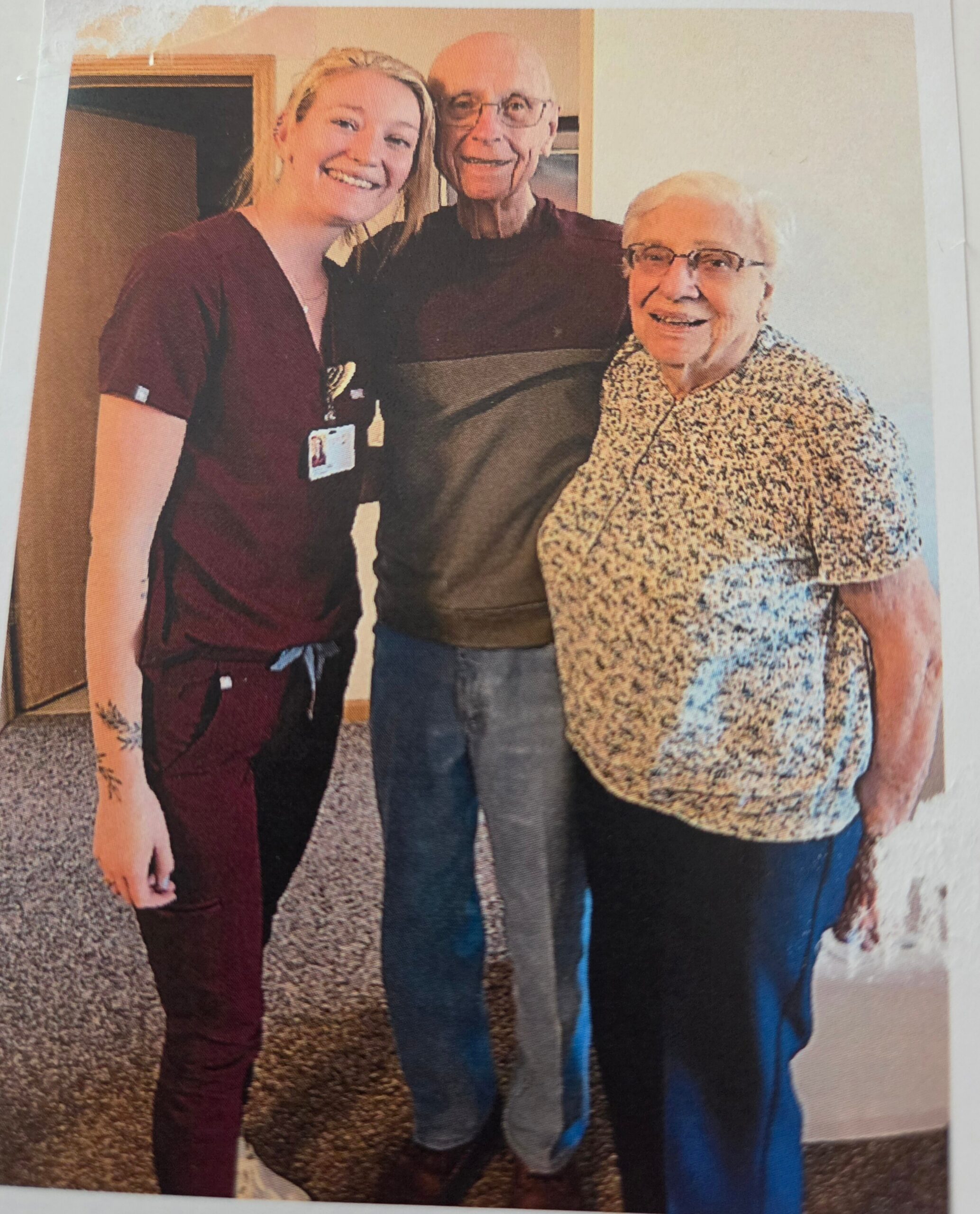 Three people posing indoors: a smiling young nurse in burgundy scrubs with an elderly man and woman beside her