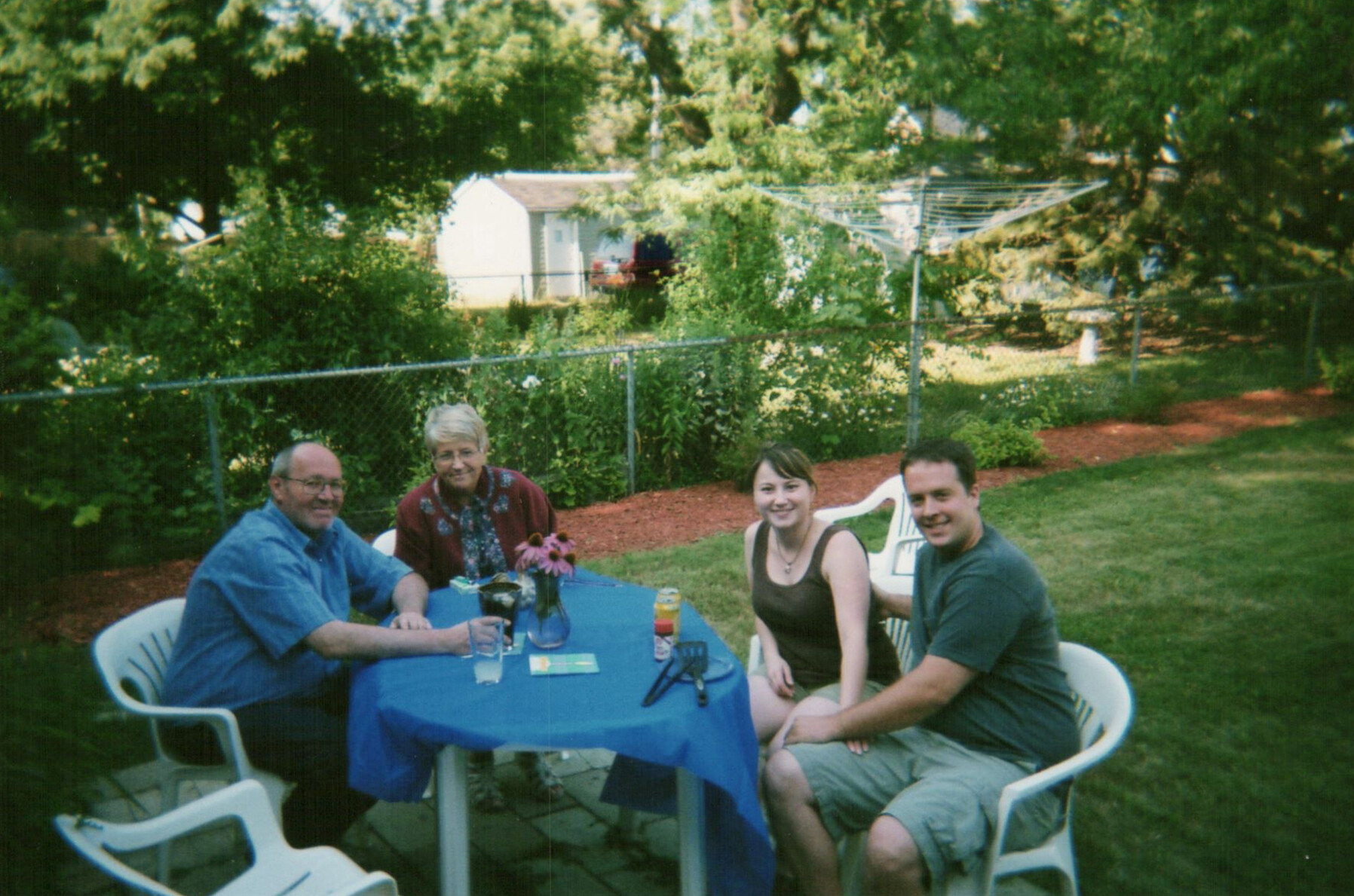 Four people sit around a blue table in a sunny backyard garden, enjoying drinks and conversation.