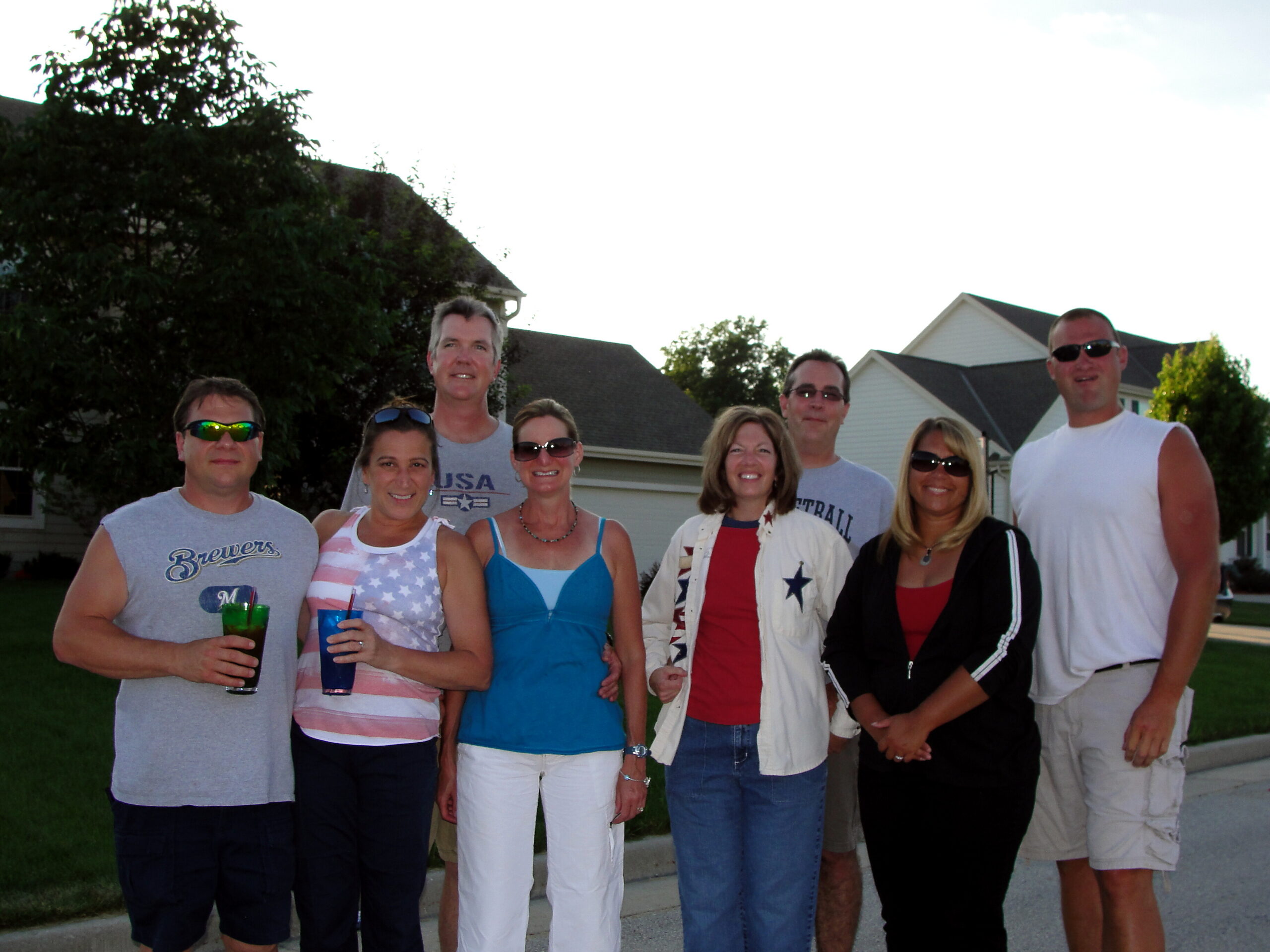 Group of adults posing together on a suburban sidewalk, smiling for the camera, holding colorful drinks (cups) and wearing casual summer clothing.