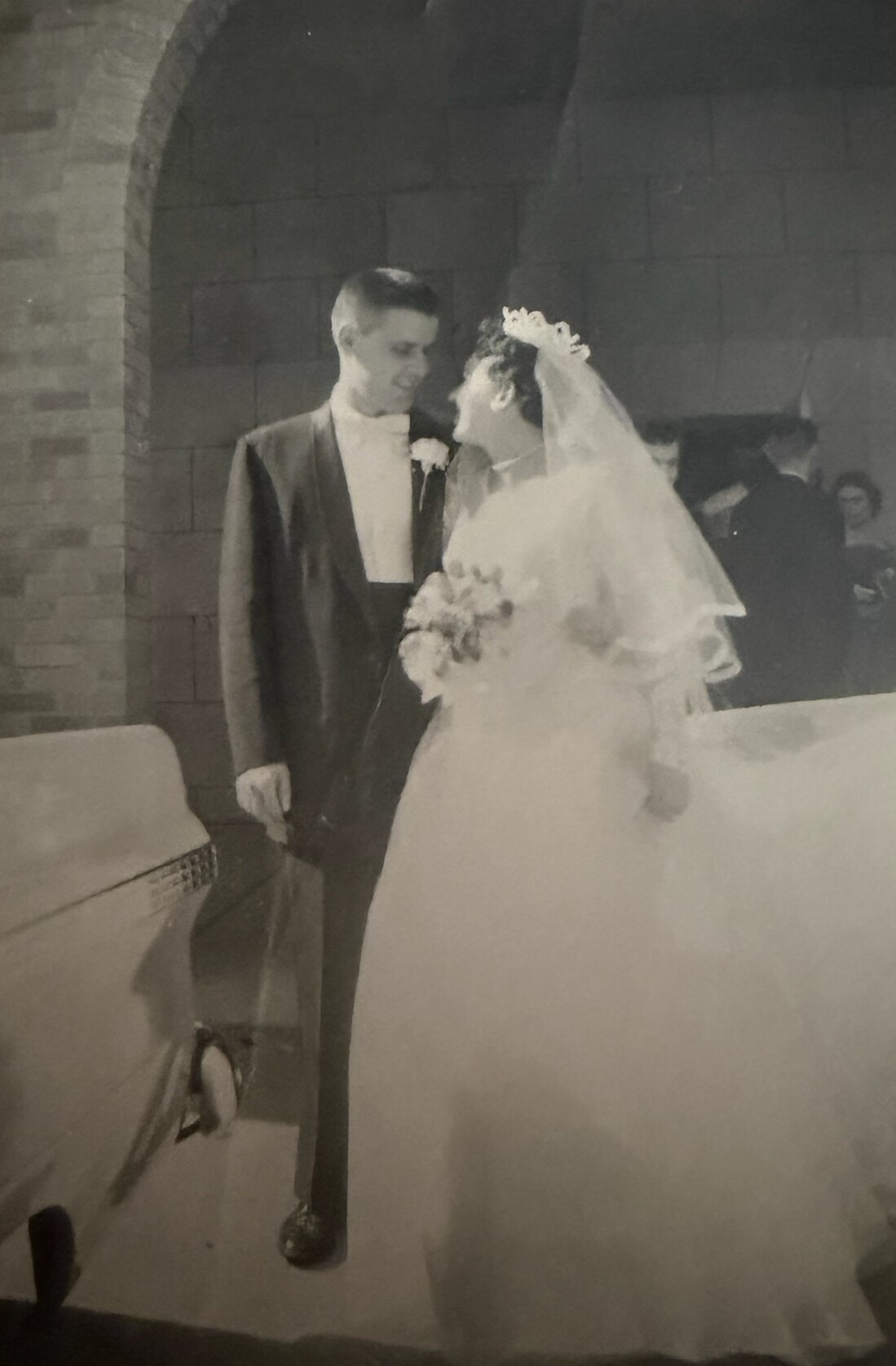 Bride and groom stand together in a wedding dress and tuxedo, smiling at each other, inside a church with an arch behind them; the bride holds a bouquet and wears a veil.
