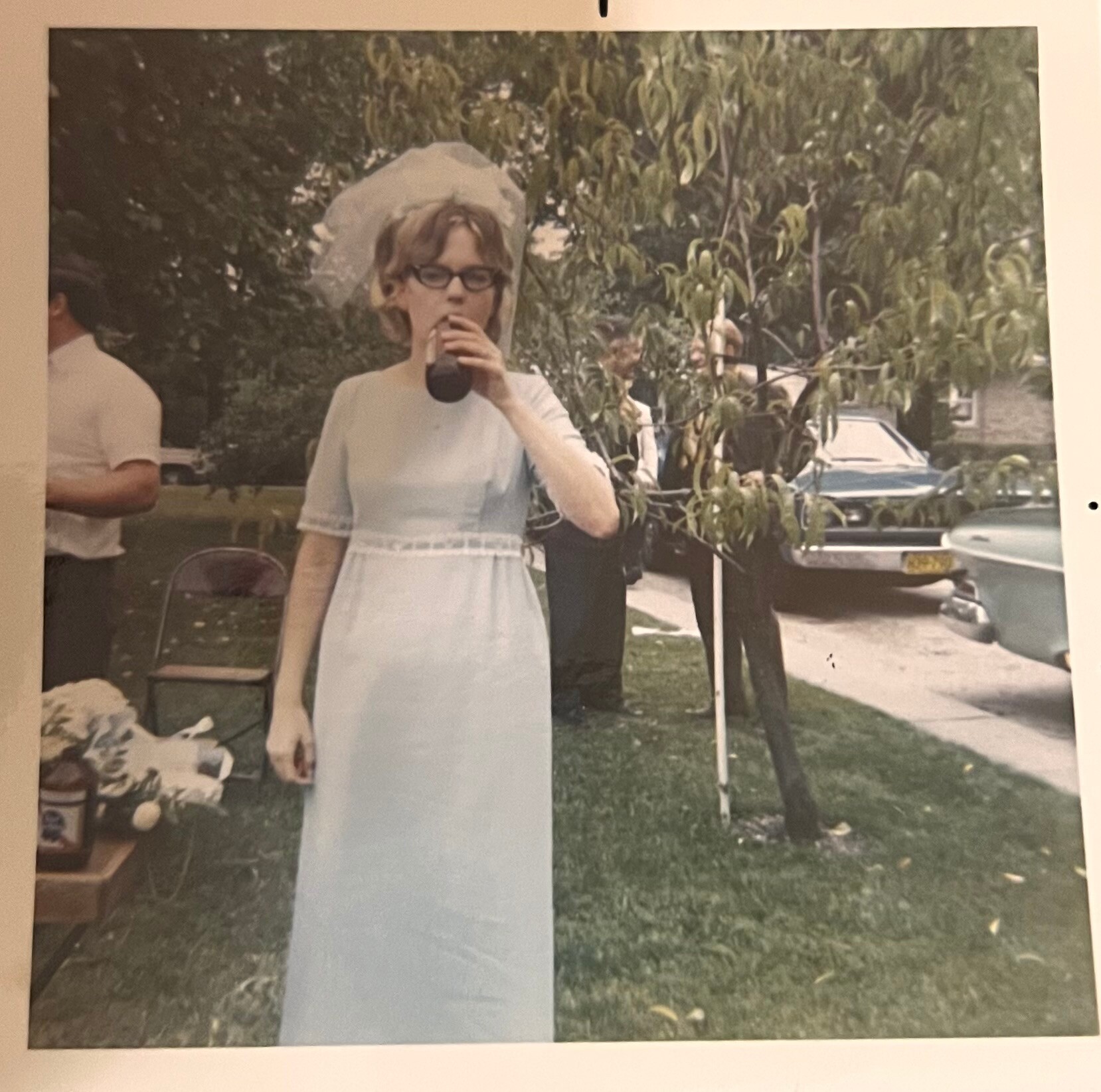 Bride wearing a light blue dress and veil, drinking from a bottle outdoors at a yard, with trees and a parked car in the background.