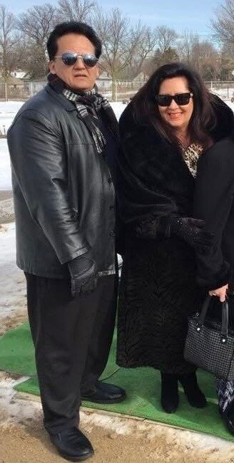 Two adults posing together outdoors in winter, wearing dark coats and sunglasses, standing on a green mat with snow in the background.