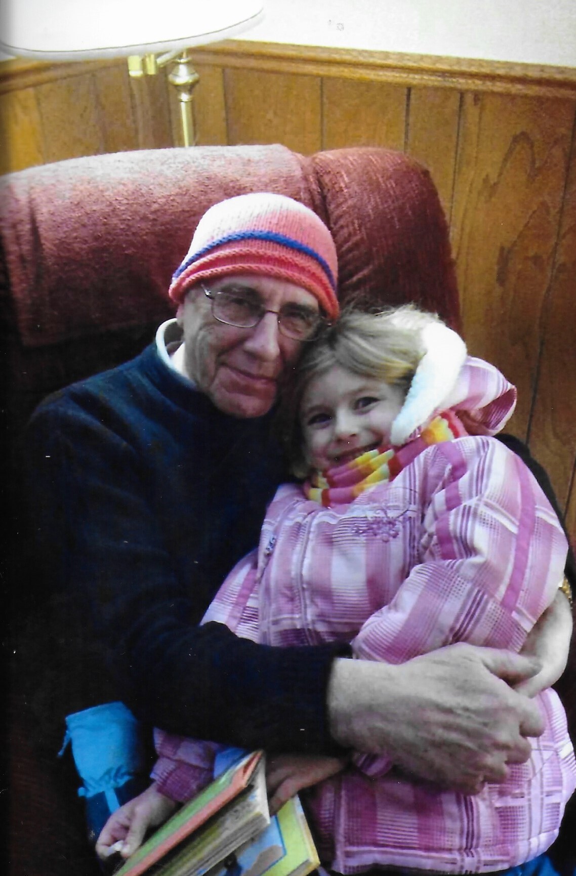 An elderly man in a red-and-pink knit hat holds a smiling young girl in a pink plaid jacket while sitting on a brown couch in a wood-panelled room.