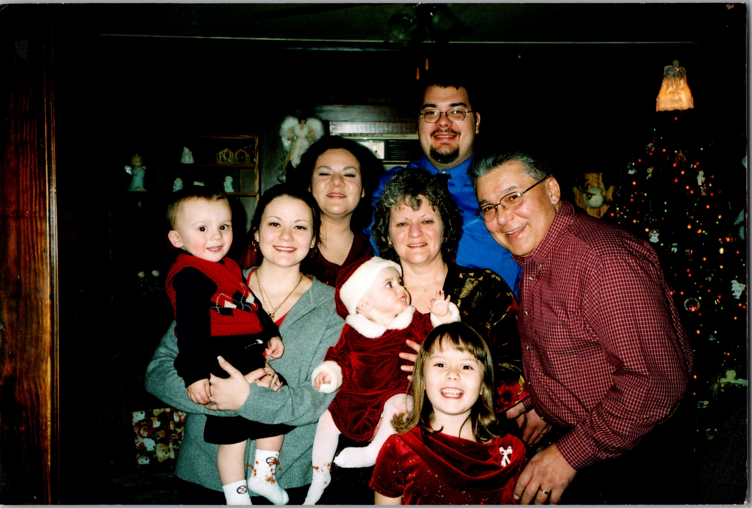 A multi-generational family smiles for a Christmas photo indoors, with a decorated tree in the background and kids in festive clothing.