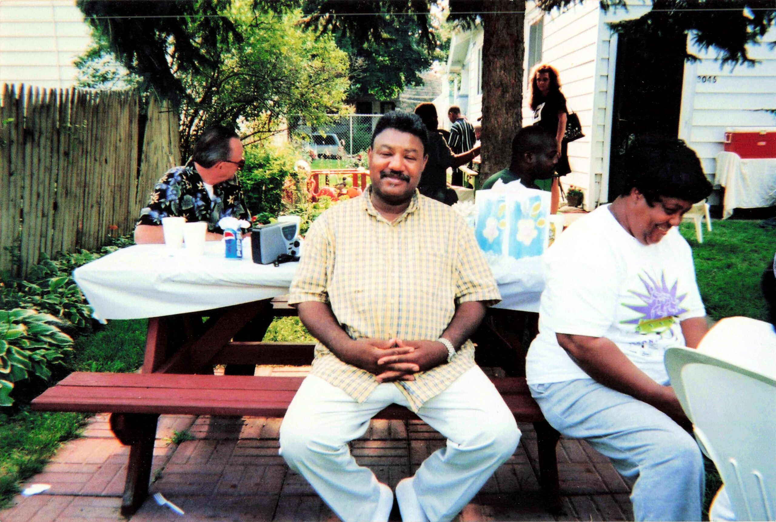 Smiling man in a yellow plaid shirt sits at a wooden picnic table in a backyard party, with people and a house in the background.