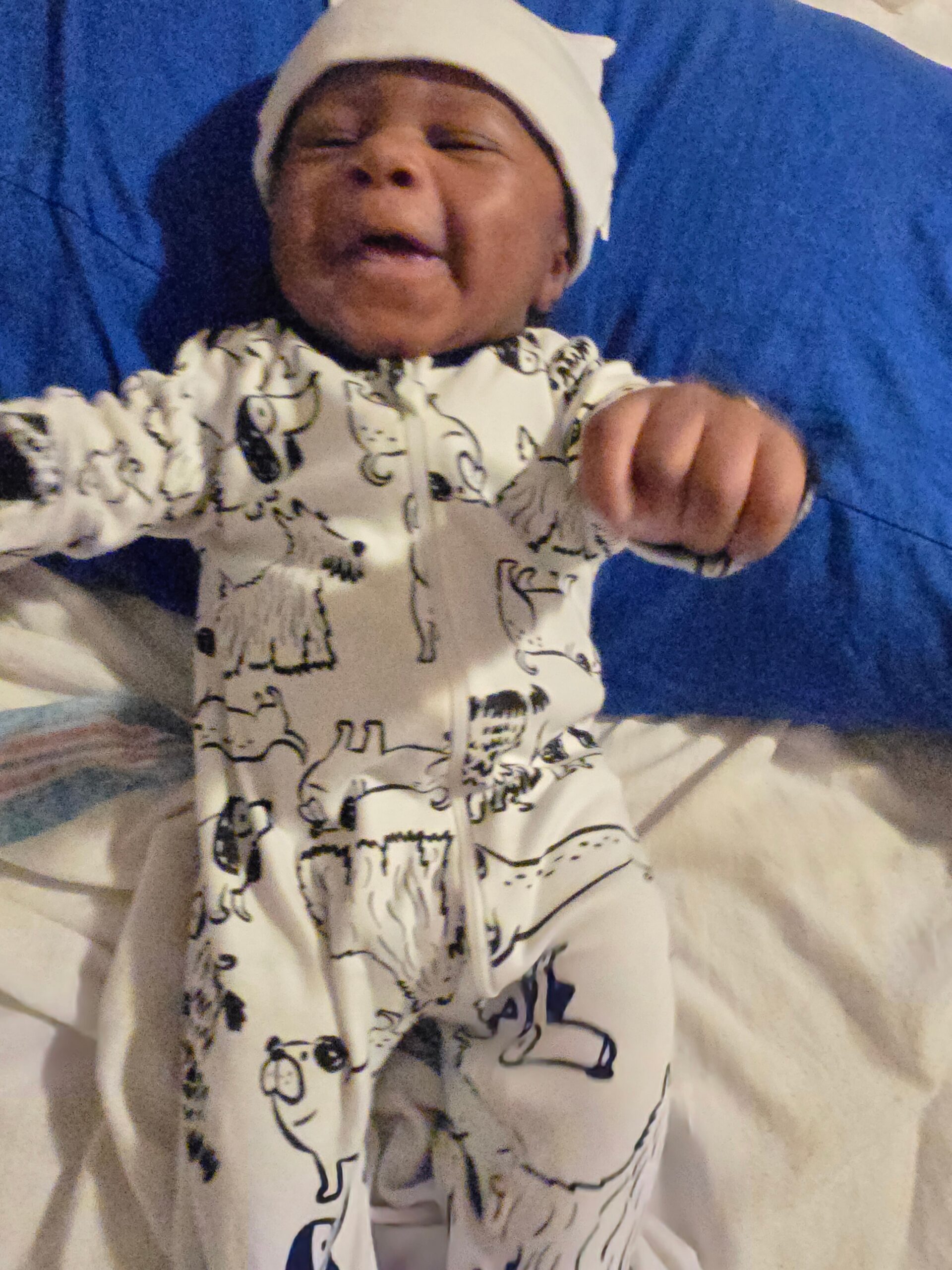 Newborn baby wearing a white hat and a patterned white outfit, lying on a blue hospital blanket with eyes closed.
