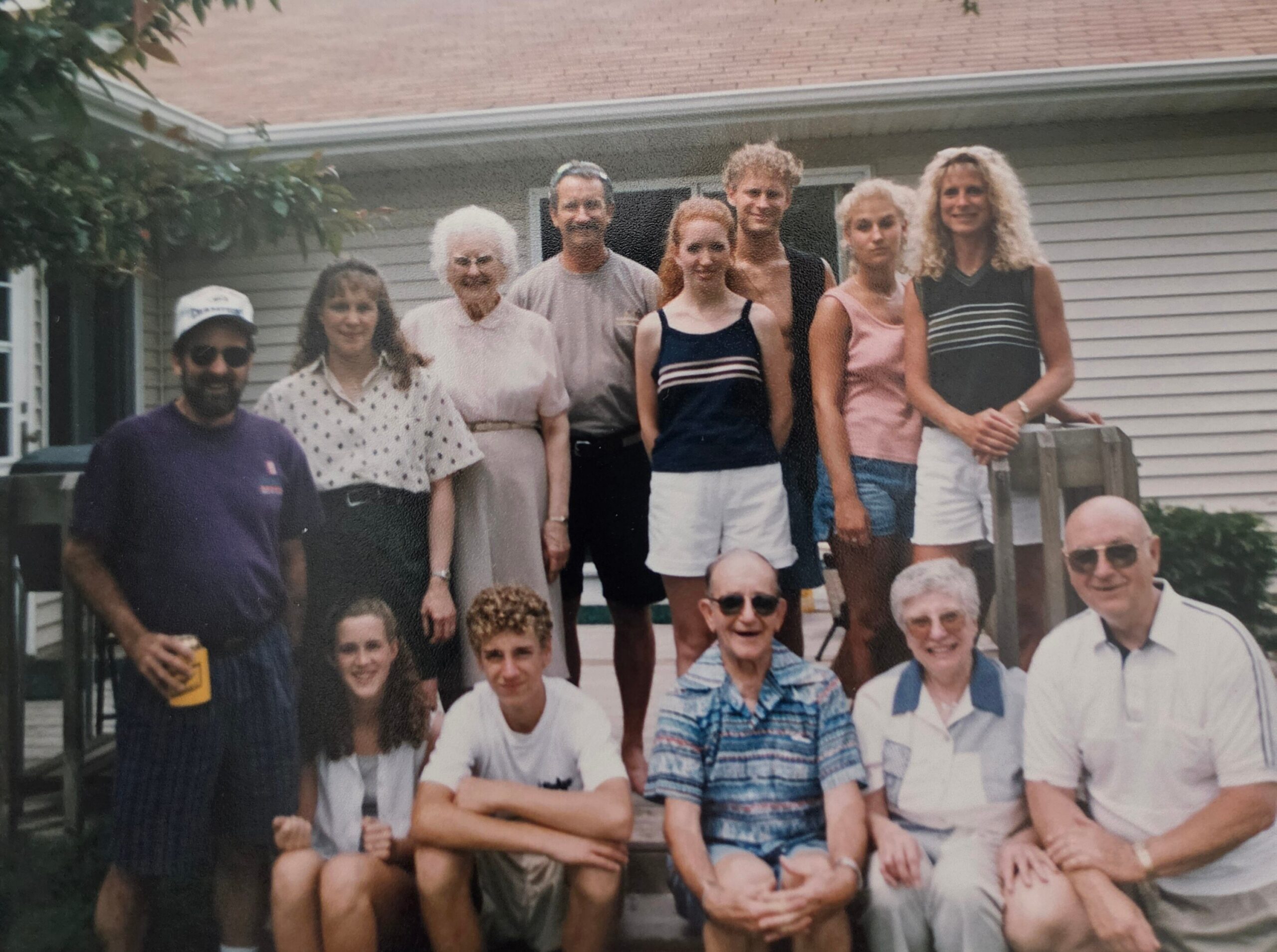 Group of adults and teens posing together on a wooden deck in front of a suburban house, summer clothing and relaxed smiles.