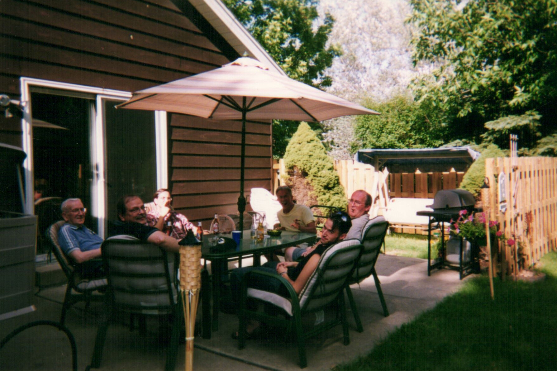 Group of people seated around a glass patio table under a beige umbrella in a sunny backyard.] ,