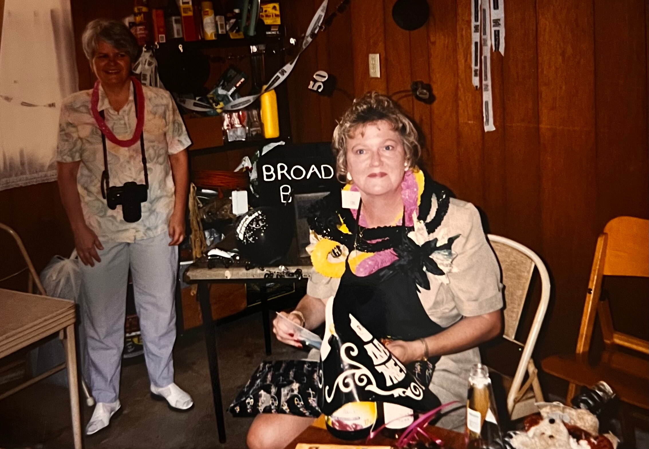 Older woman with a colorful scarf seated at a table, wearing a black-and-yellow patterned outfit, as another woman stands nearby with a camera around her neck in a cluttered room decorated for a party.