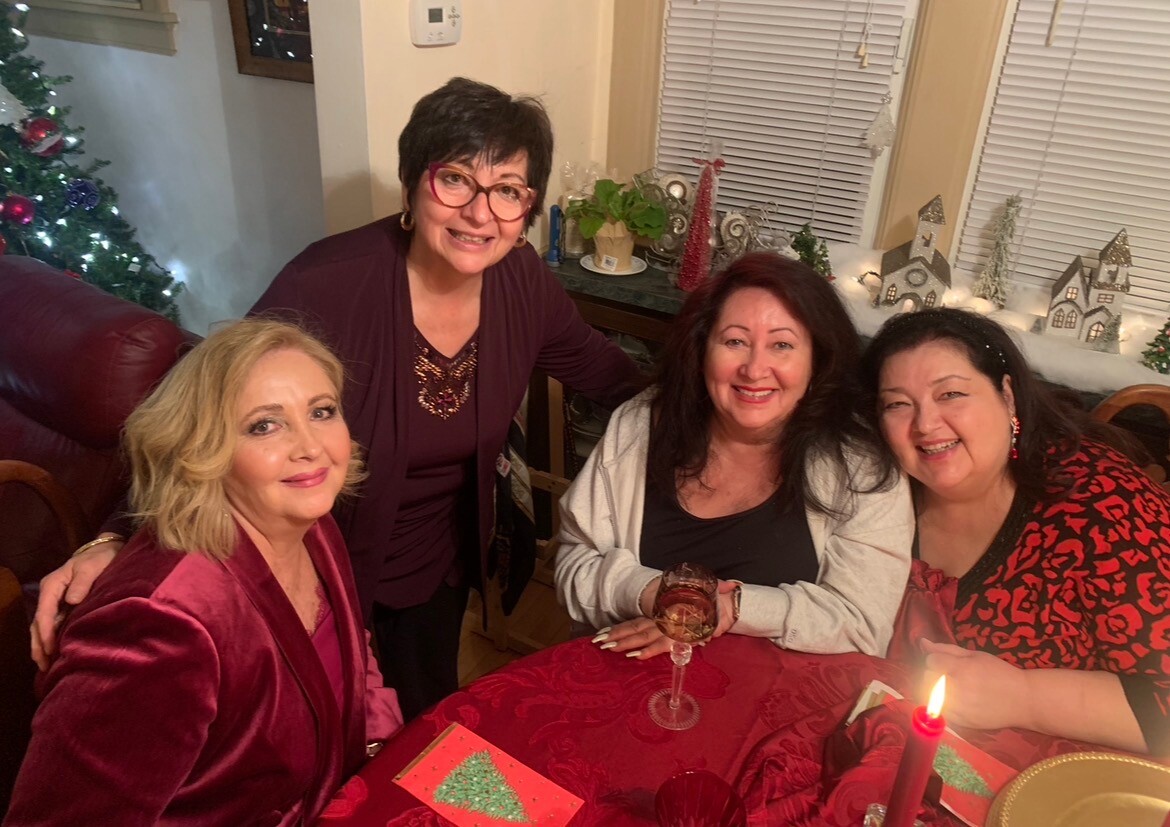 Four women smile around a red tablecloth Christmas gathering with a decorated tree in the background.