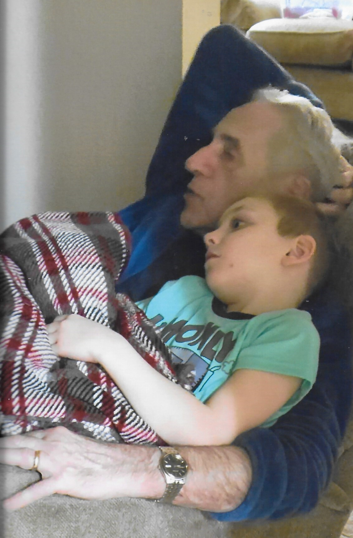 A man with gray hair sits on a couch, holding a young child wrapped in a red and gray plaid blanket.