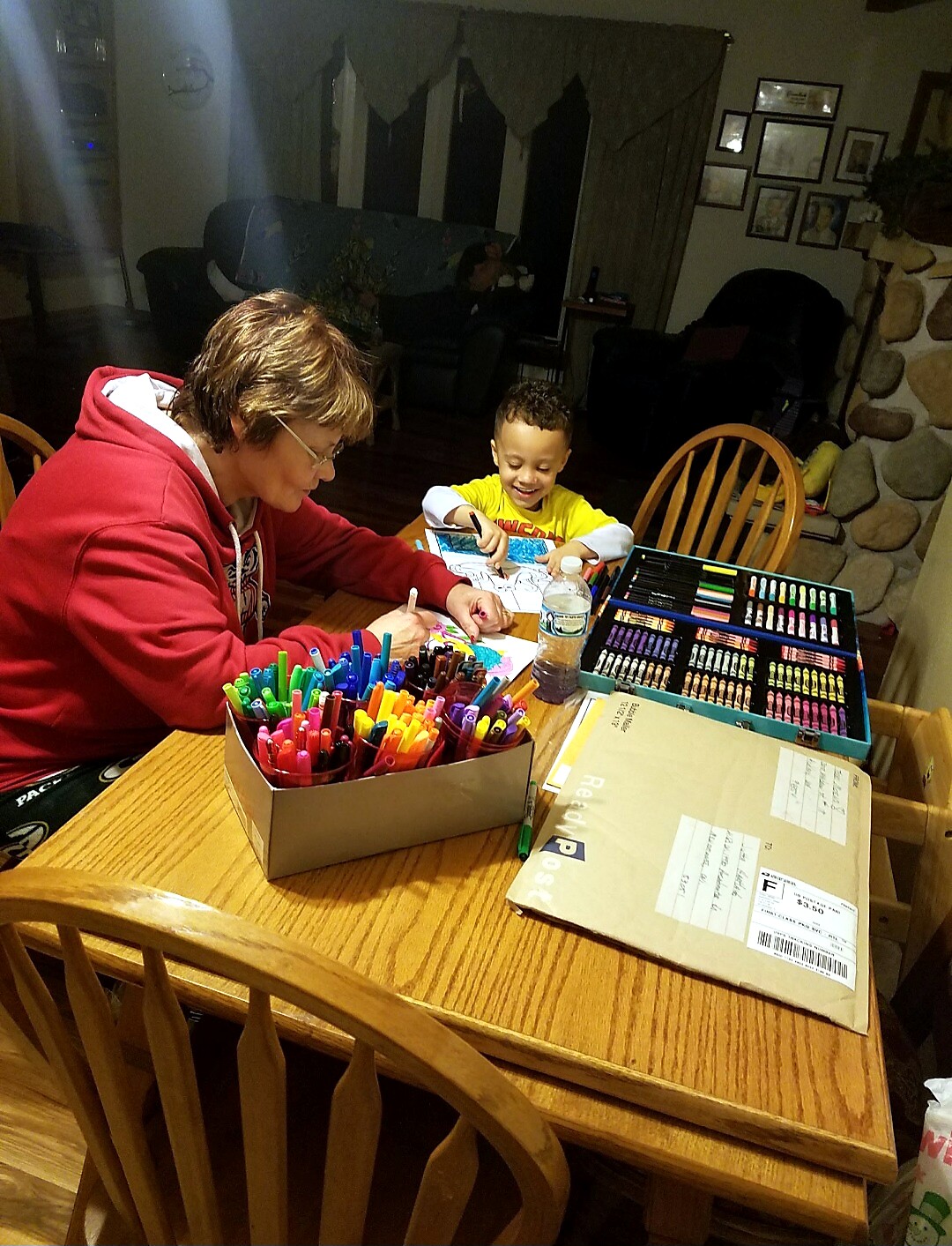 Woman in a red hoodie helping a young boy color at a dining table with markers and coloring supplies nearby.