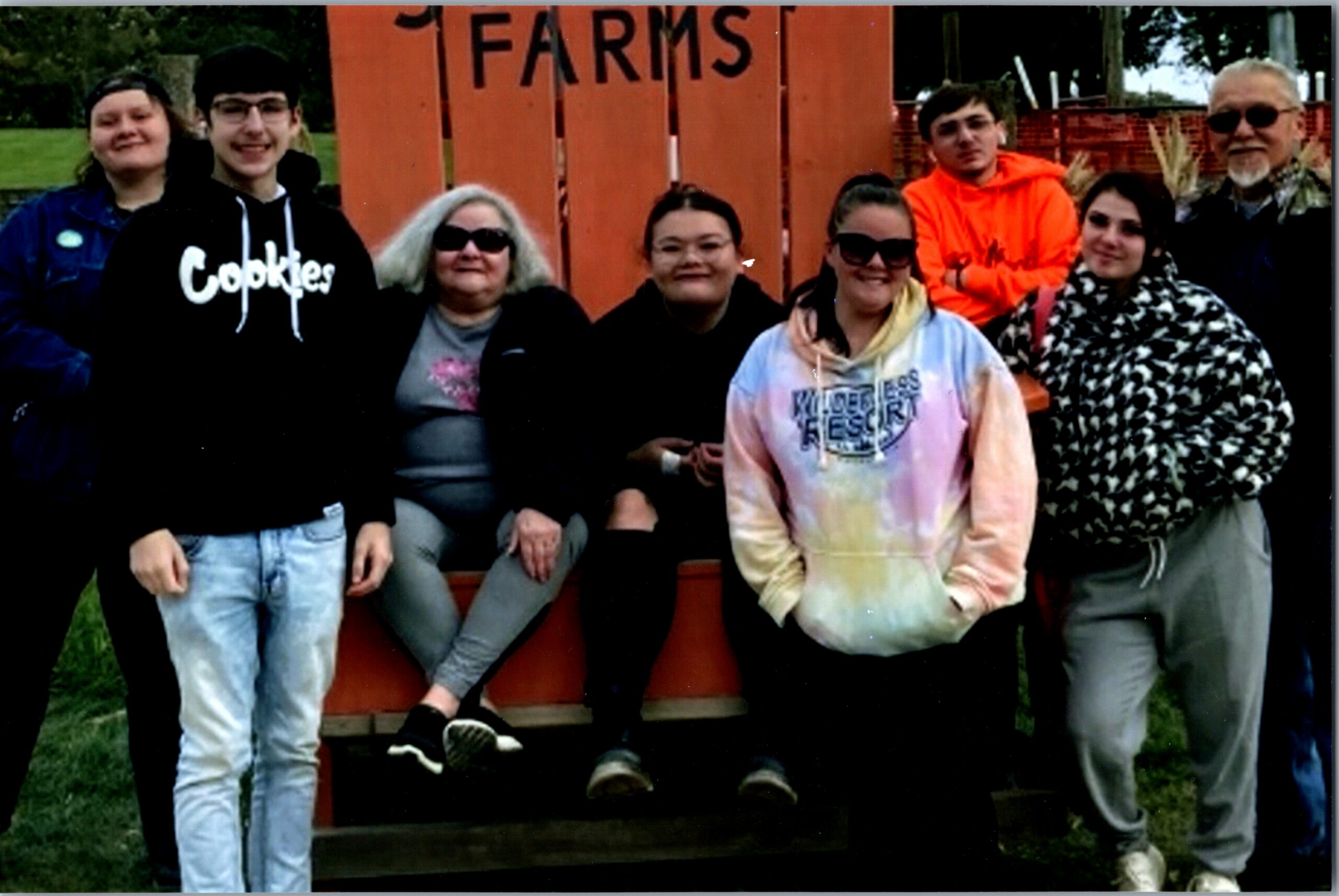 Group of eight people posing together outdoors in front of an orange wooden fence. They wear casual clothing and sunglasses on a cloudy day.