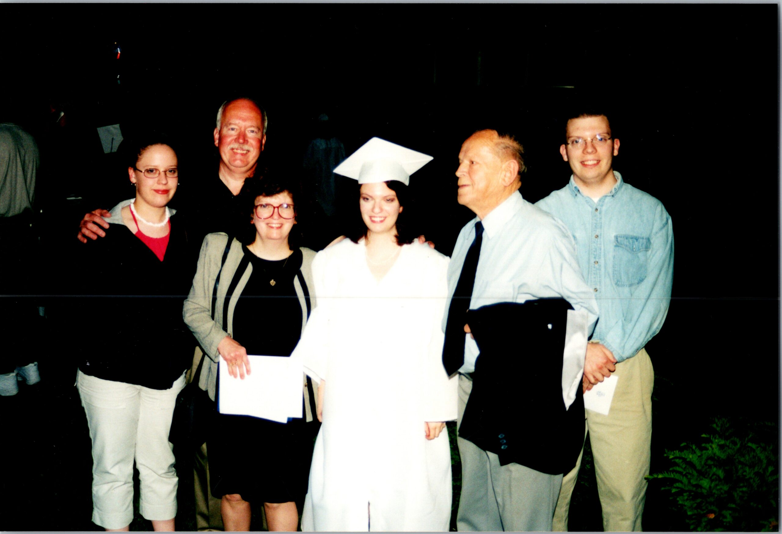 Group of six adults posing together outdoors at night, with a graduate in a white cap and gown in the center smiling holding a diploma.