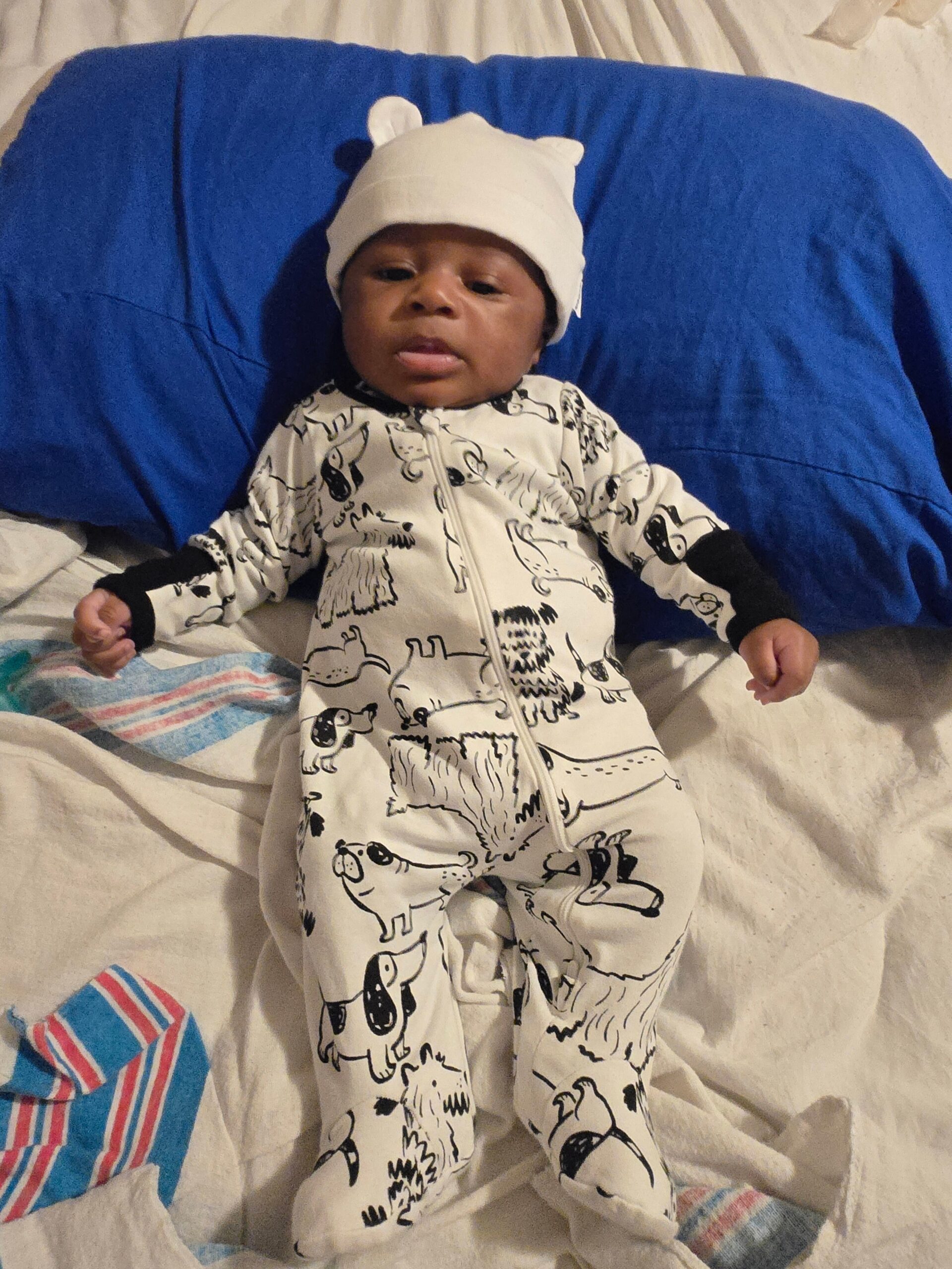 Baby lying on a bed wearing a white hat with small ears and a white onesie with black animal prints against a blue blanket.