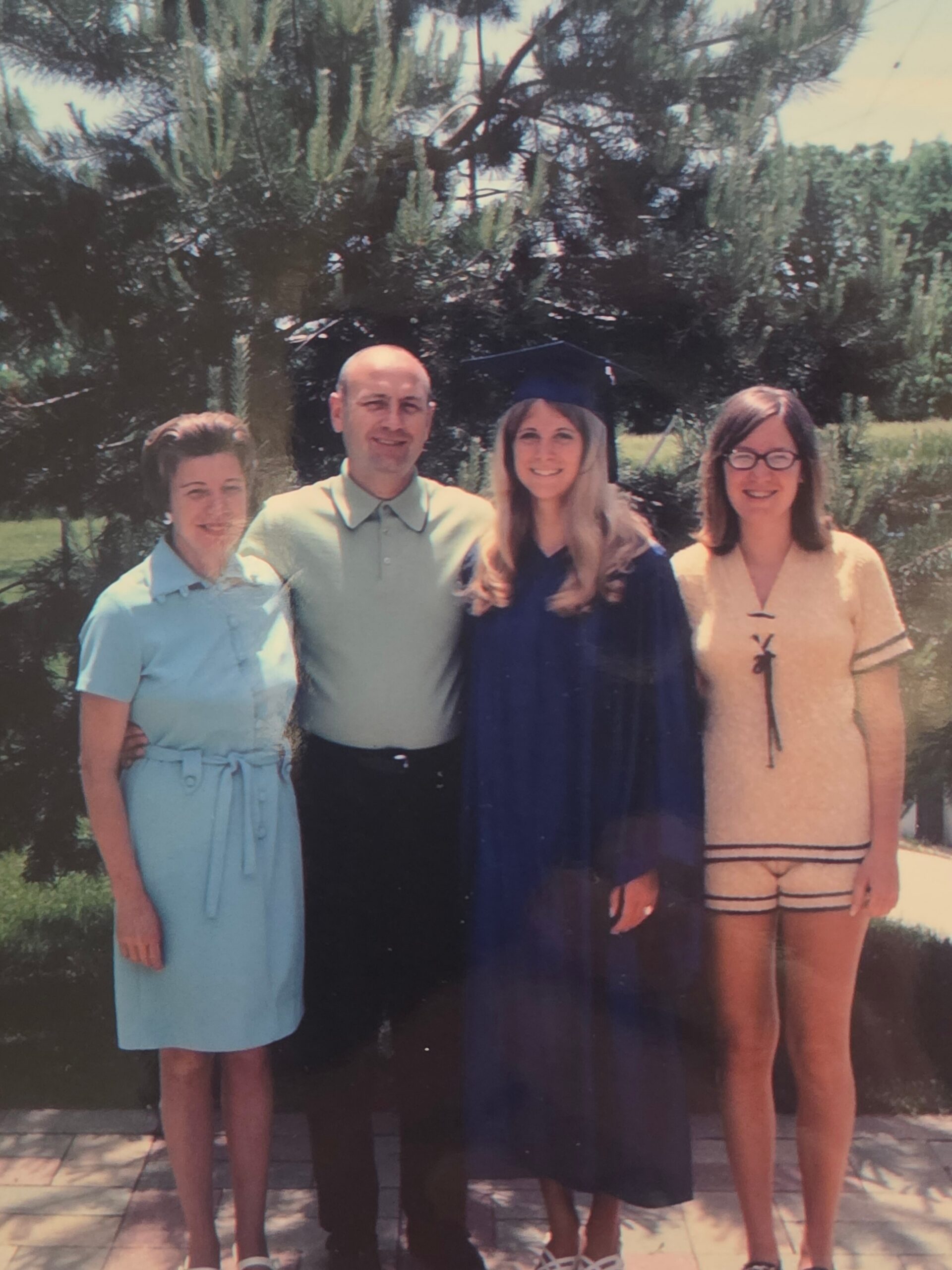 Group photo outdoors with four people; a graduate in a blue gown and cap stands with three relatives against trees in a sunny yard.