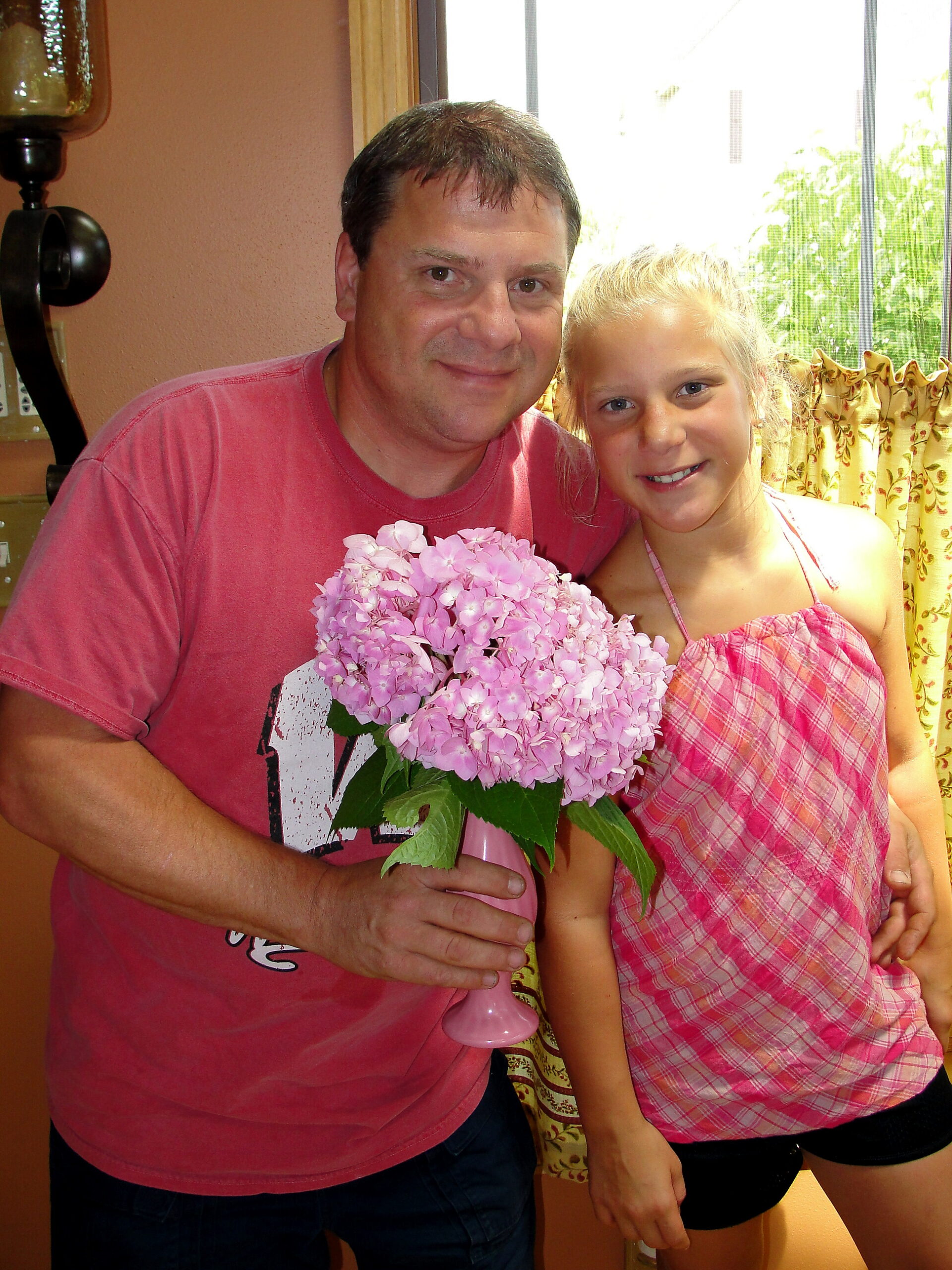 Man and girl smiling while holding a pink hydrangea bouquet indoors by a windowed wall.
