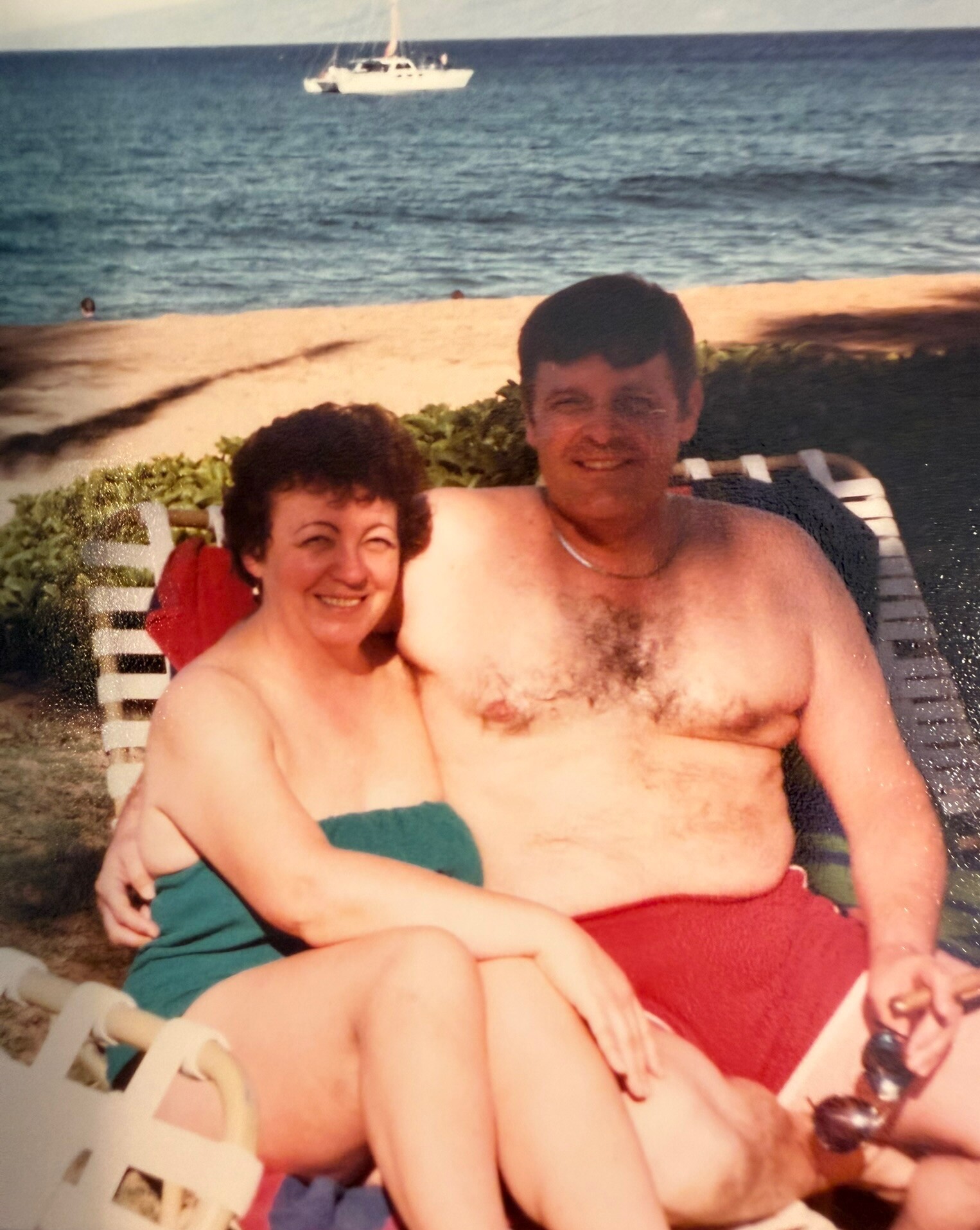 Smiling couple sits on a beach chair by the sea;Woman wears teal swimsuit, man is shirtless in red shorts with a towel nearby; boat in the distant water.