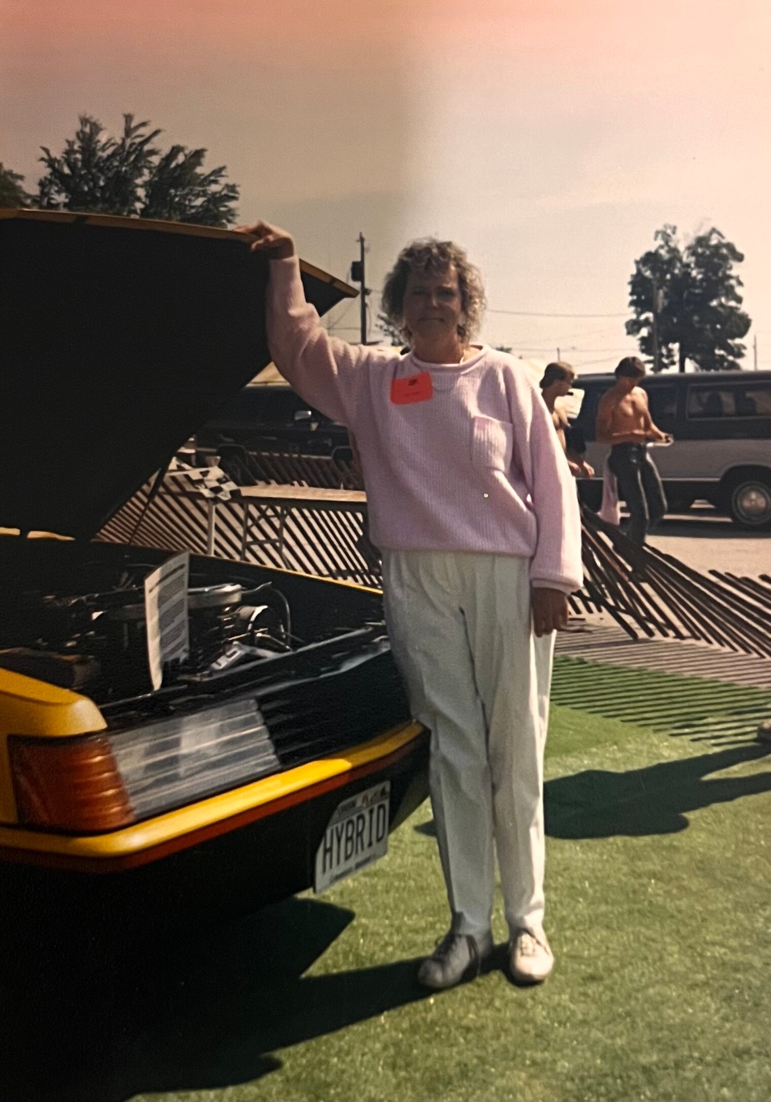 Woman with curly hair in a pink sweater stands beside an open car trunk at an outdoor car show; badge on sweater and white pants visible.