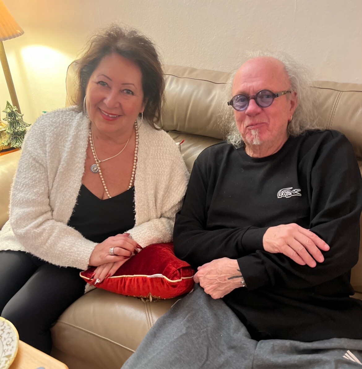 Smiling woman and man sit closely on a beige couch in a cozy living room, the woman holding a red decorative pillow between them.