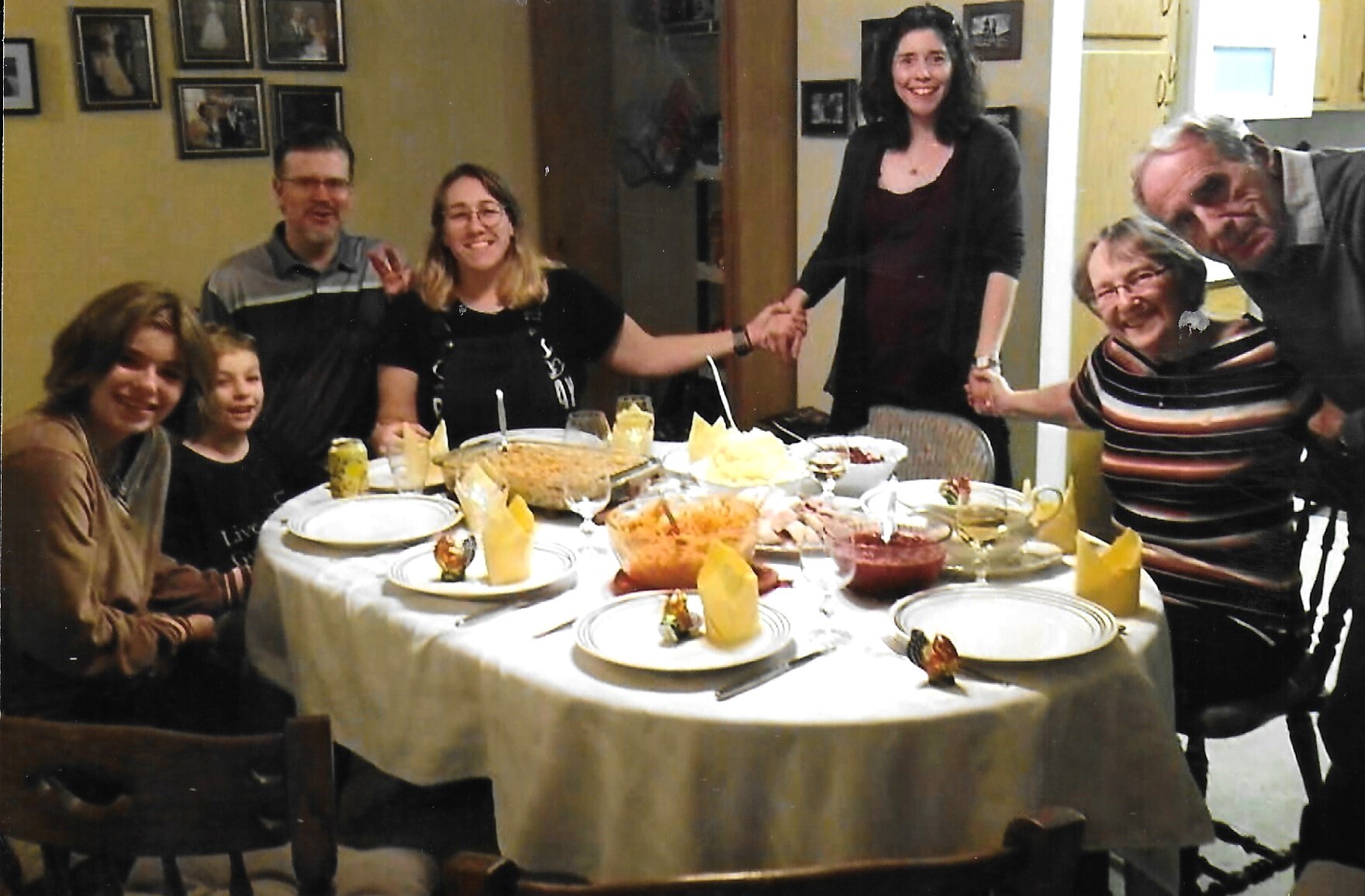A multigenerational family sits around a dining table, smiling as they hold hands across the table during a meal.