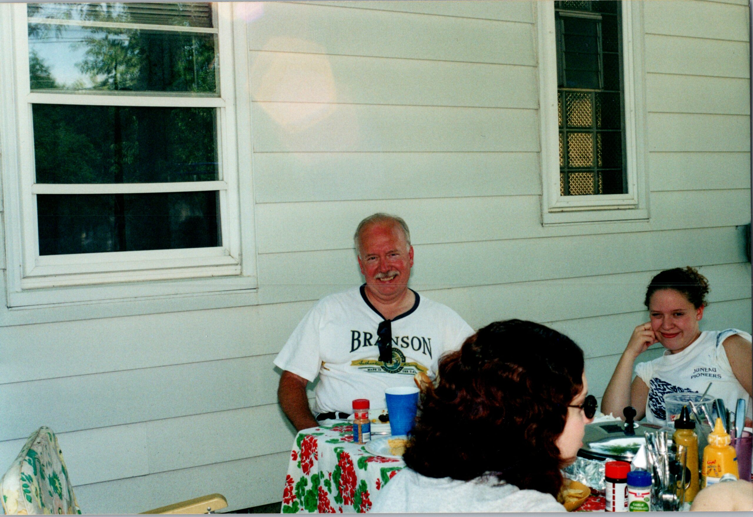 Group of people seated at a picnic table outside a light-colored house, chatting and eating together; man in a Branson shirt smiles at the camera.