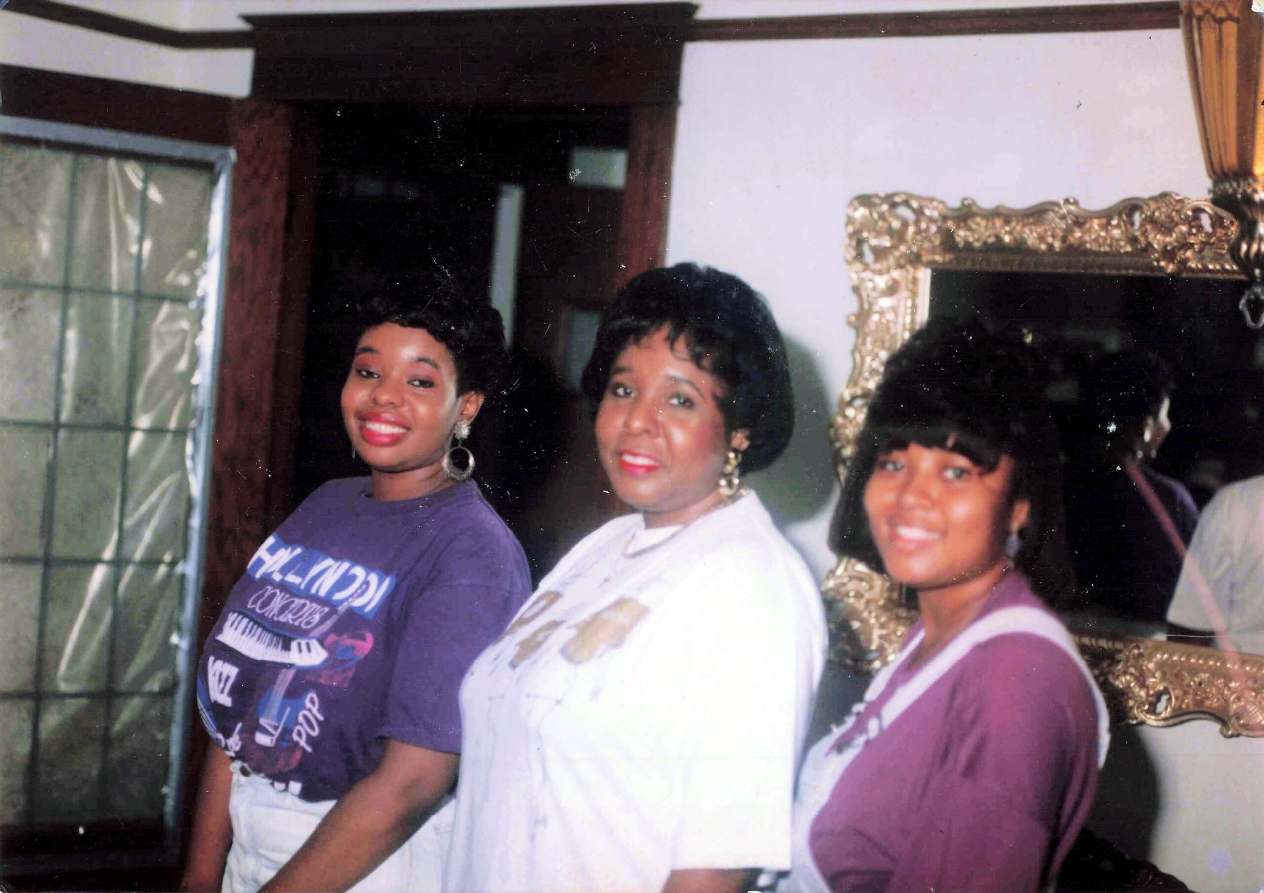 Three women pose together indoors, smiling at the camera; left wears a purple shirt, middle wears a white outfit, right wears a purple cardigan, with a decorative mirror behind them.