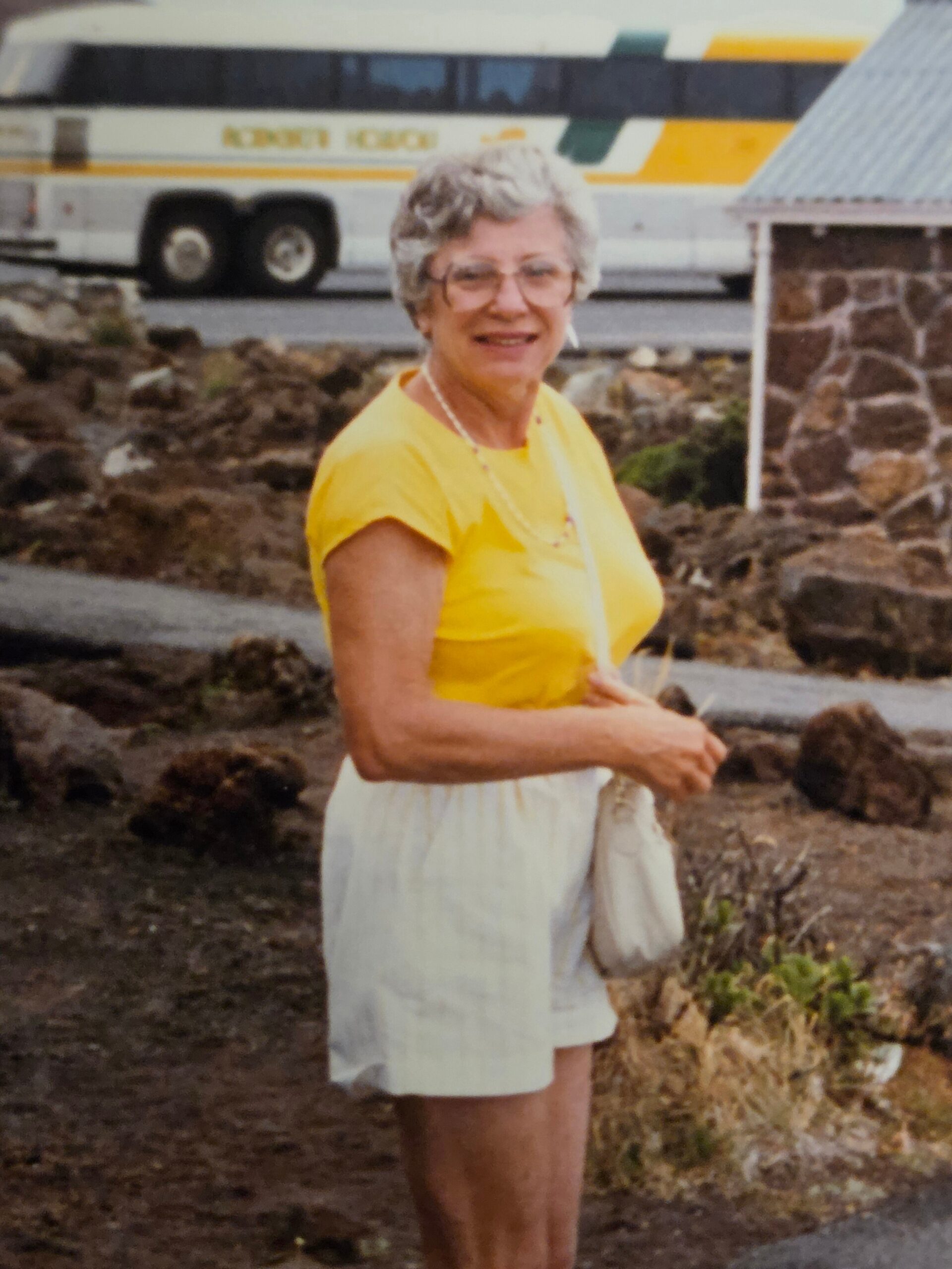Older woman in a yellow top and light shorts standing outdoors, smiling, with a large bus in the background and rocky ground nearby.