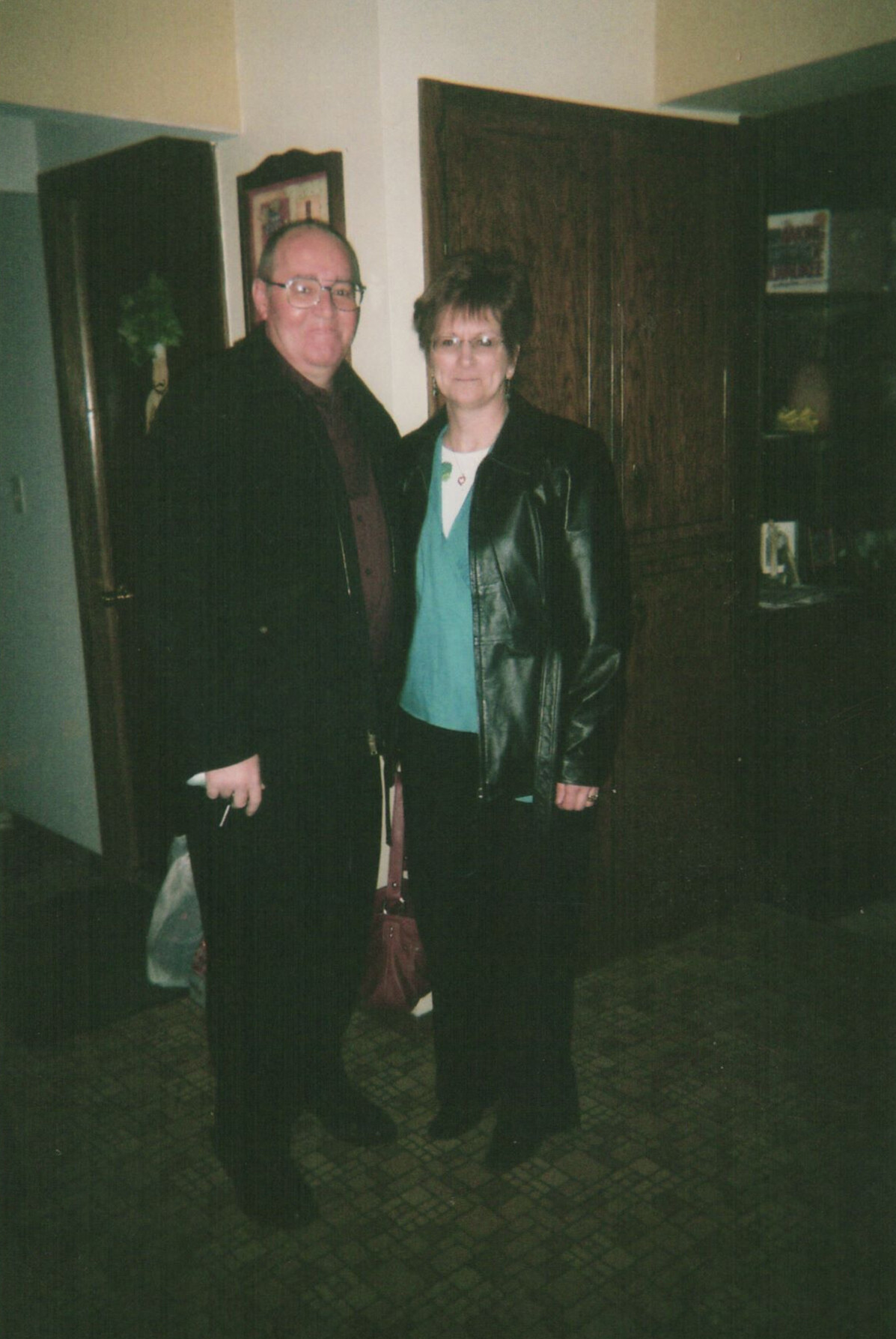 A man and a woman stand together in a dim hallway, both wearing dark jackets and glasses, facing the camera.