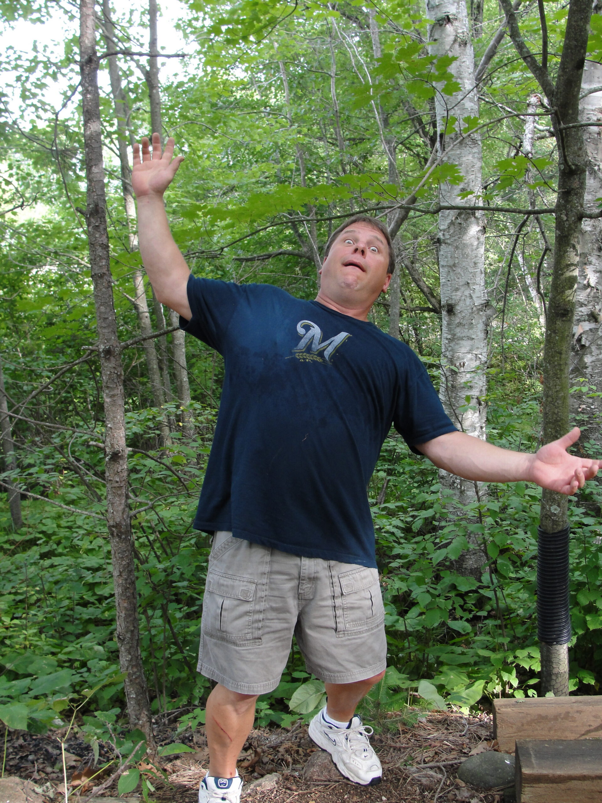 Man in a blue T-shirt and tan shorts stands in a forest with arms raised and a surprised expression.