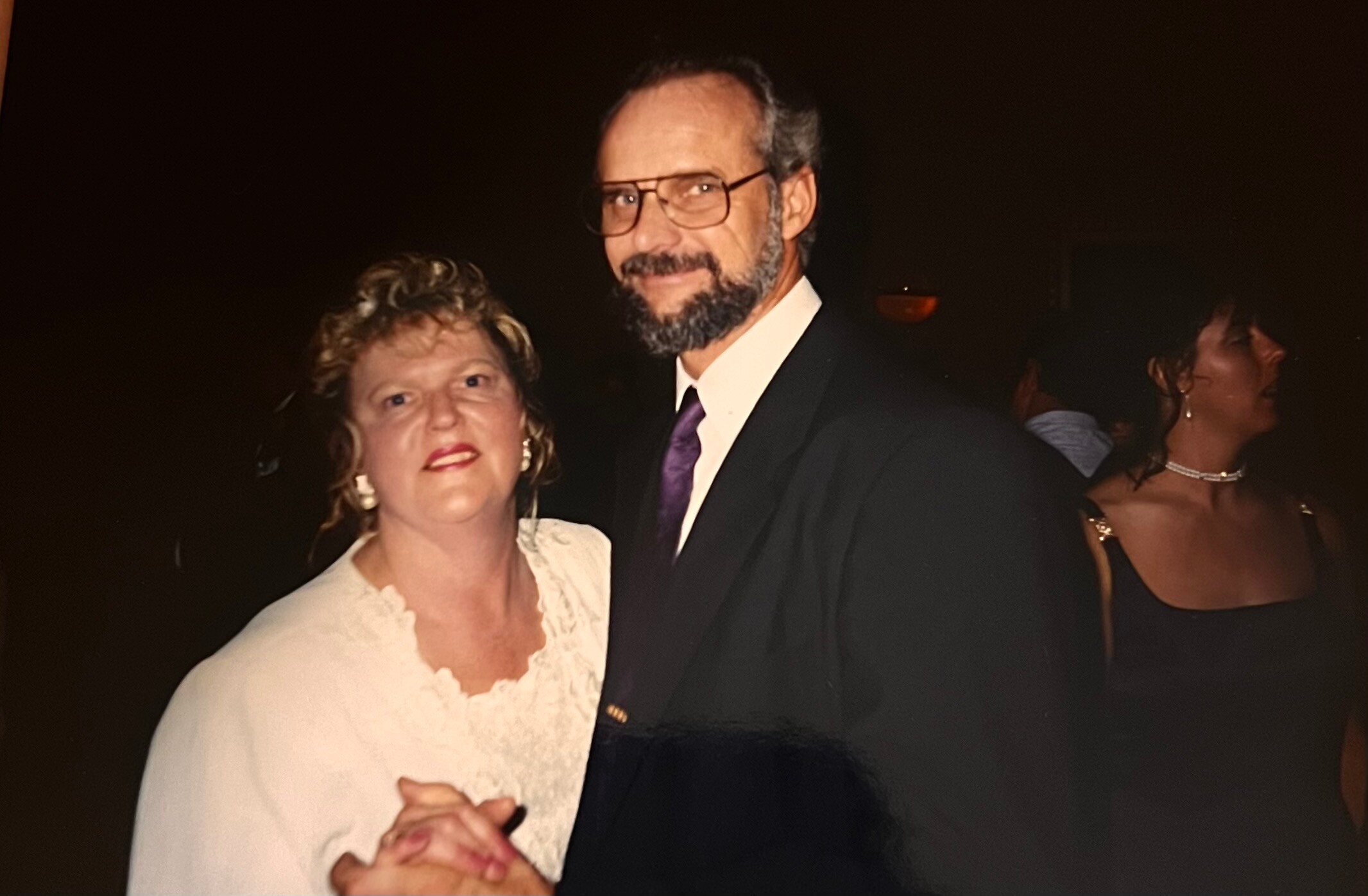 Smiling couple dancing at a formal event: man in a suit with glasses and beard beside a woman in white, dark background