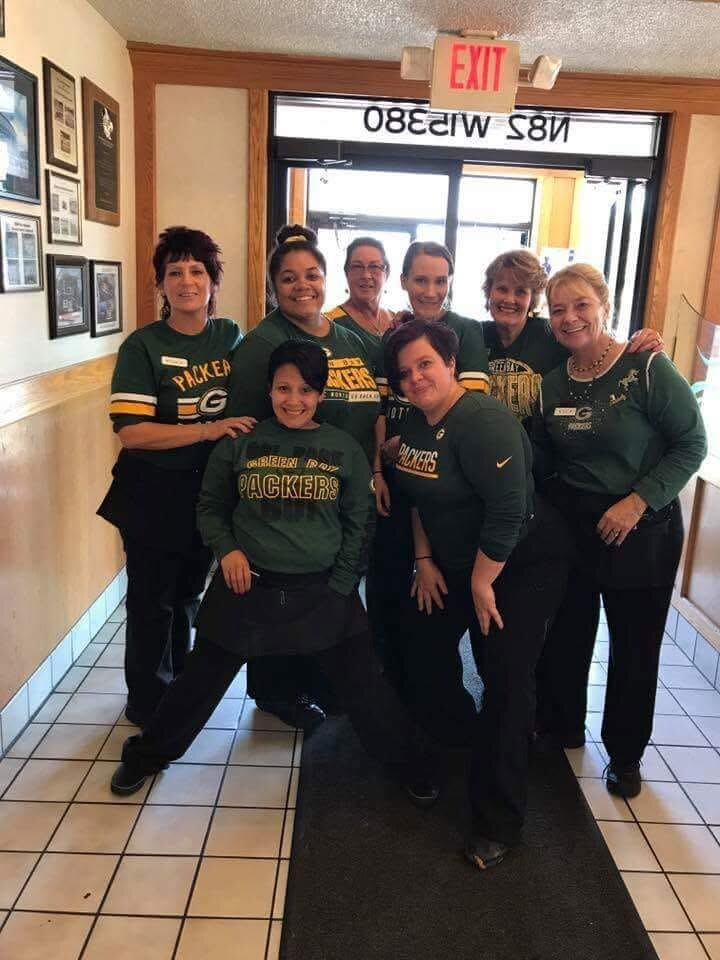 Eight women in green Packers shirts posing together in a hallway near an exit sign.
