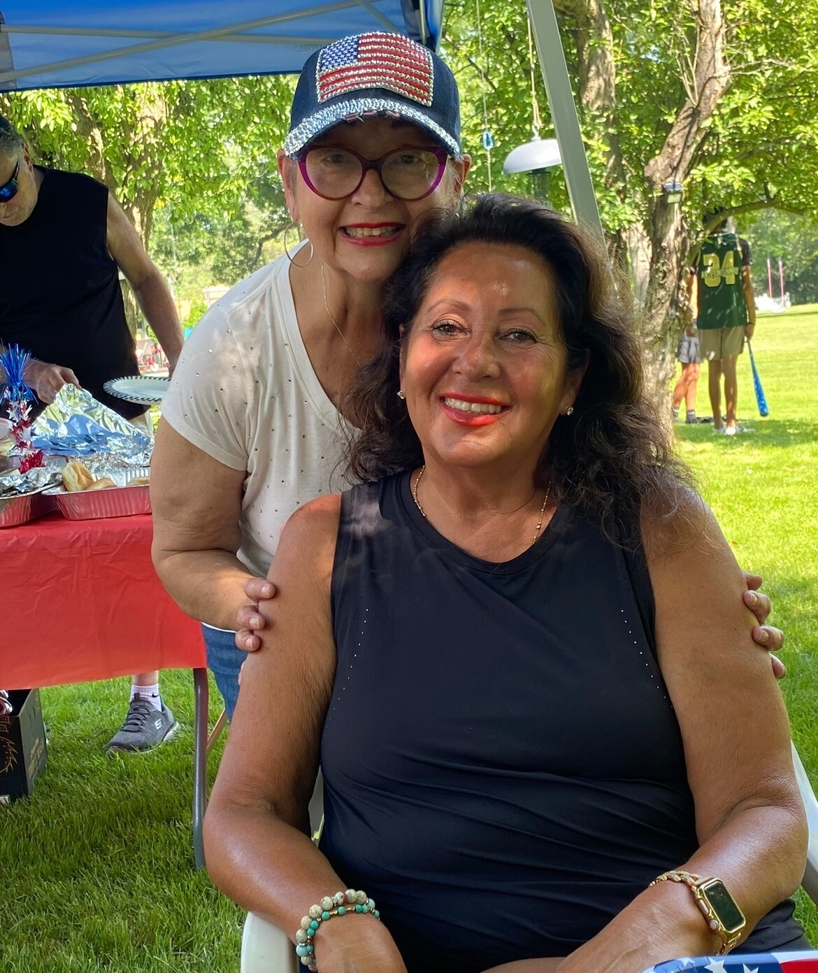 Two women smile for a photo at an outdoor park picnic, one seated in a black sleeveless top while the other wraps an arm around her from behind under a canopy.