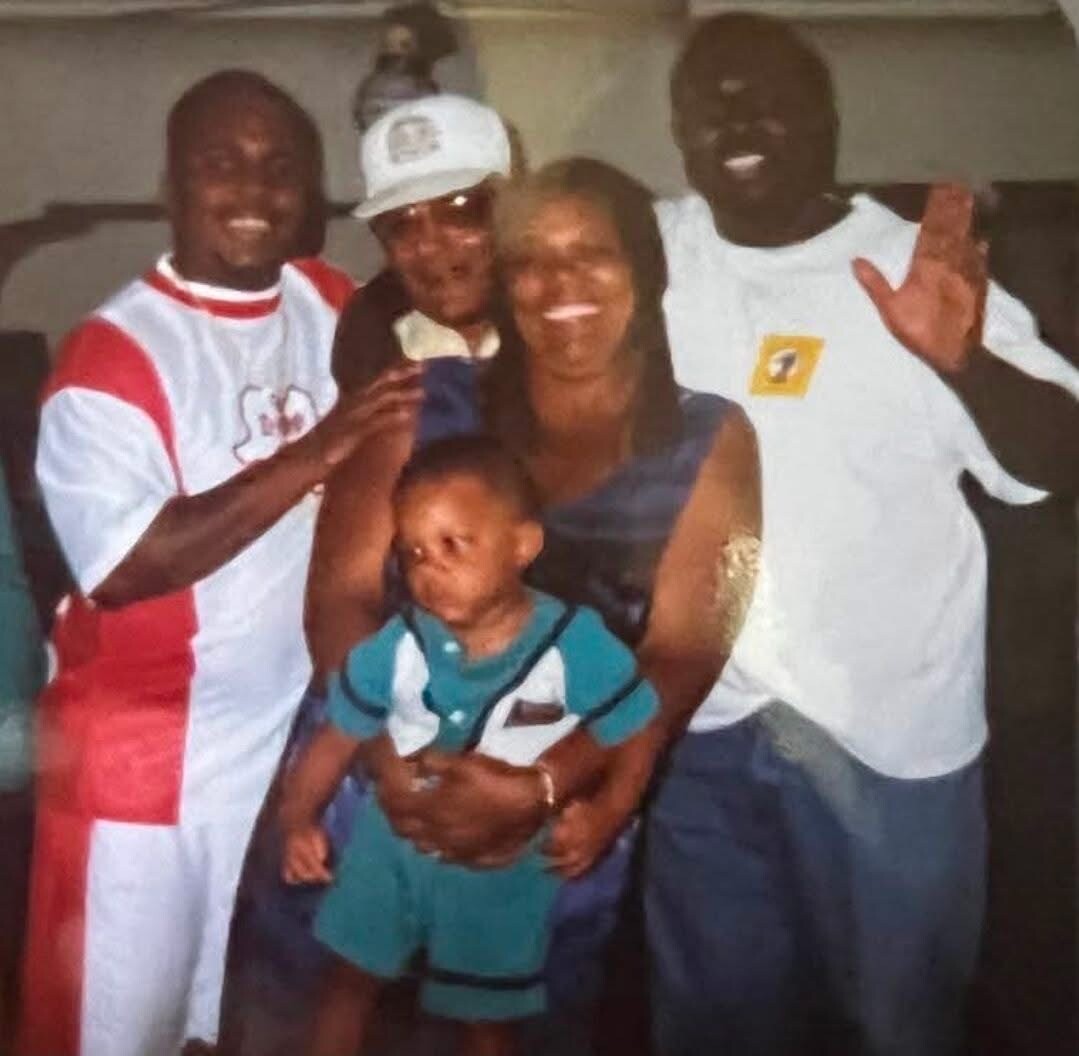Smiling family group posing for a photo: a woman holding a young boy, surrounded by three men.