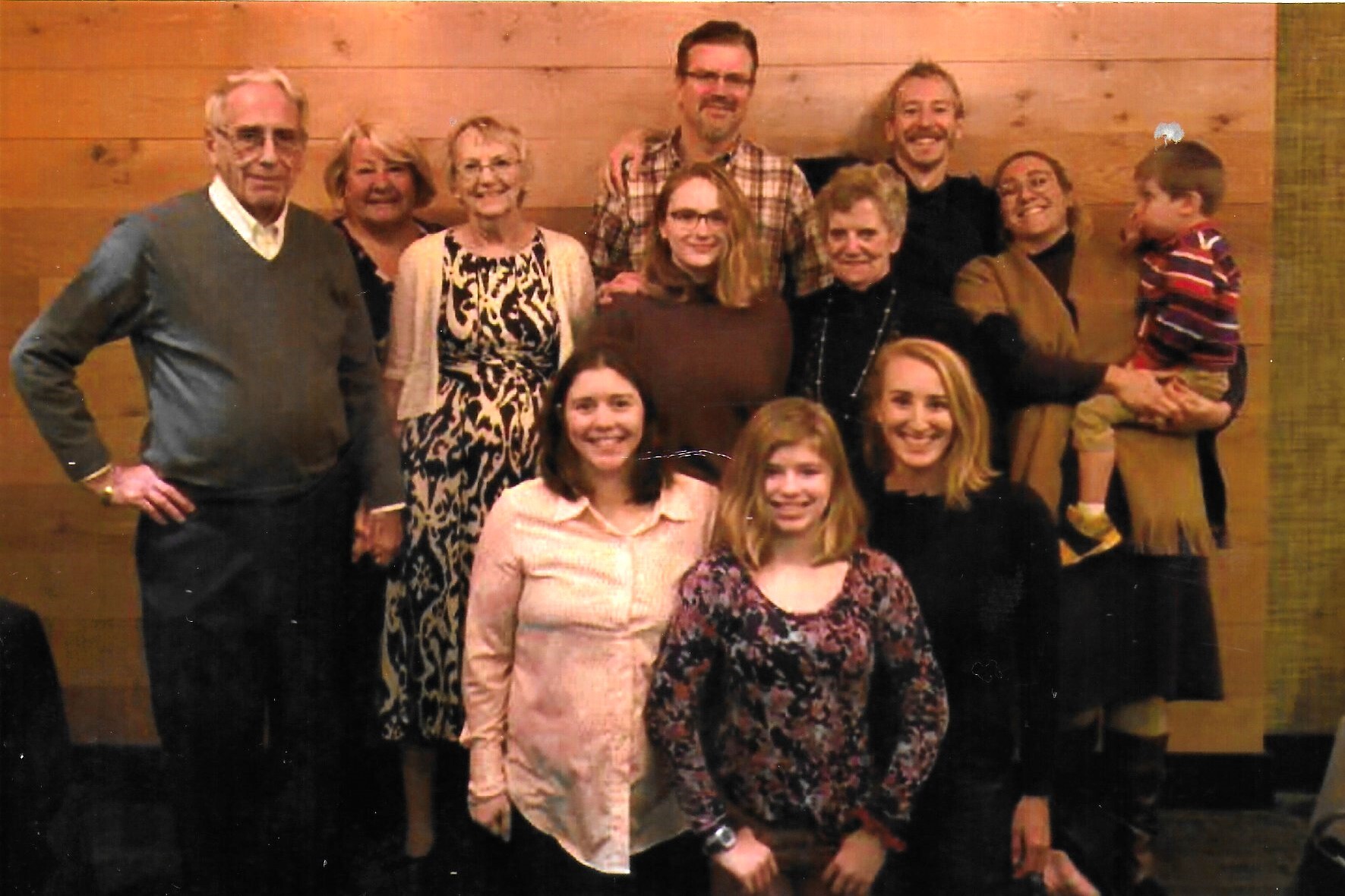 Group portrait of eleven people, adults and a child, posing indoors against a wood-paneled wall, all smiling at the camera.
