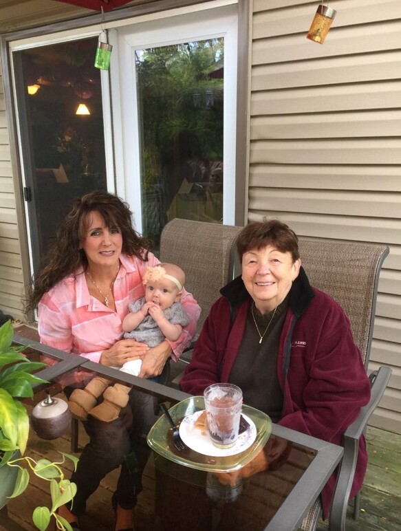 Three generations sit on a patio sofa: mom with baby and grandmother, smiling outdoors by a sliding door.
