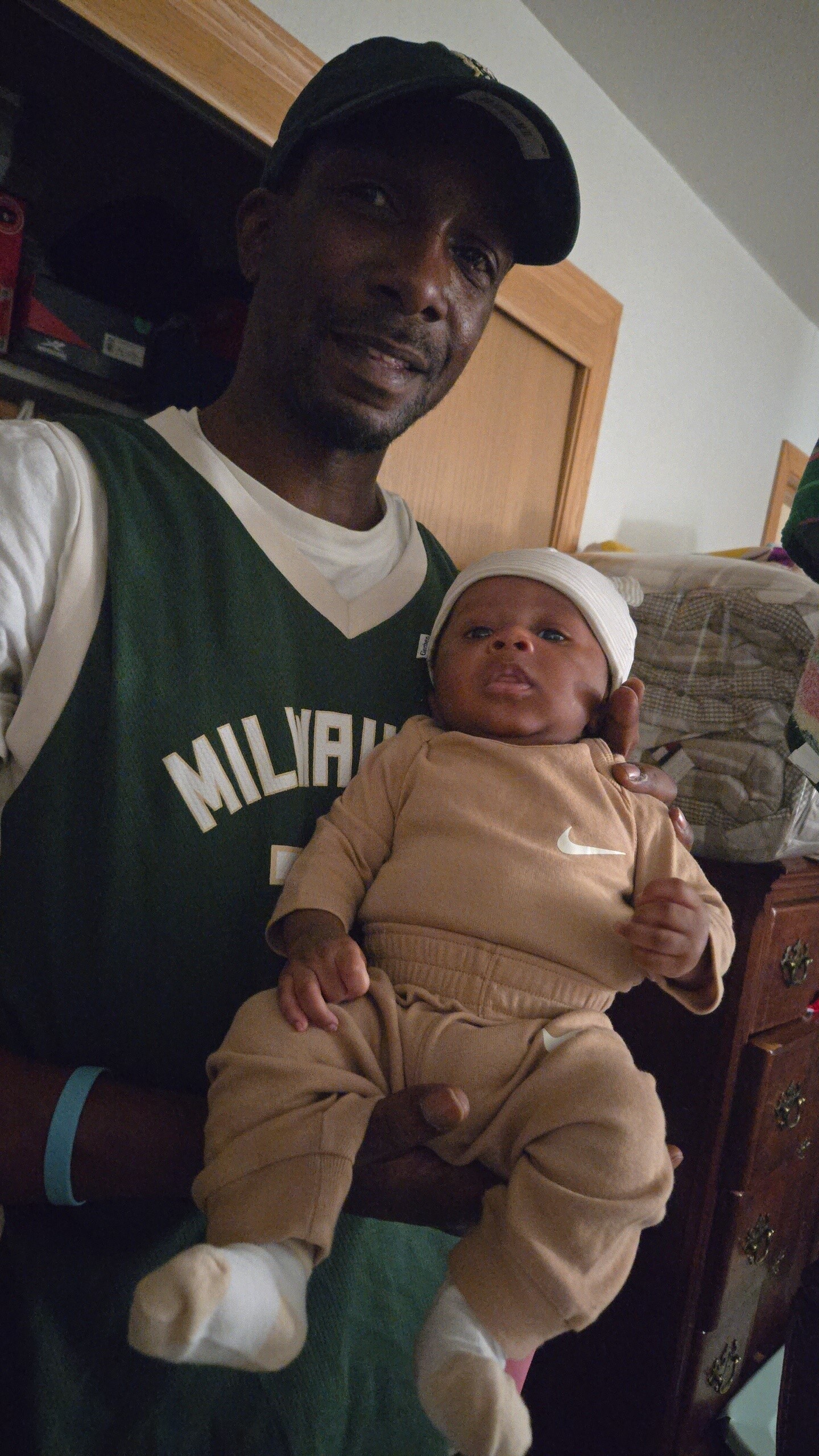 A man in a green Milwaukee jersey holds a baby dressed in beige with a white knit cap, smiling at the camera indoors.