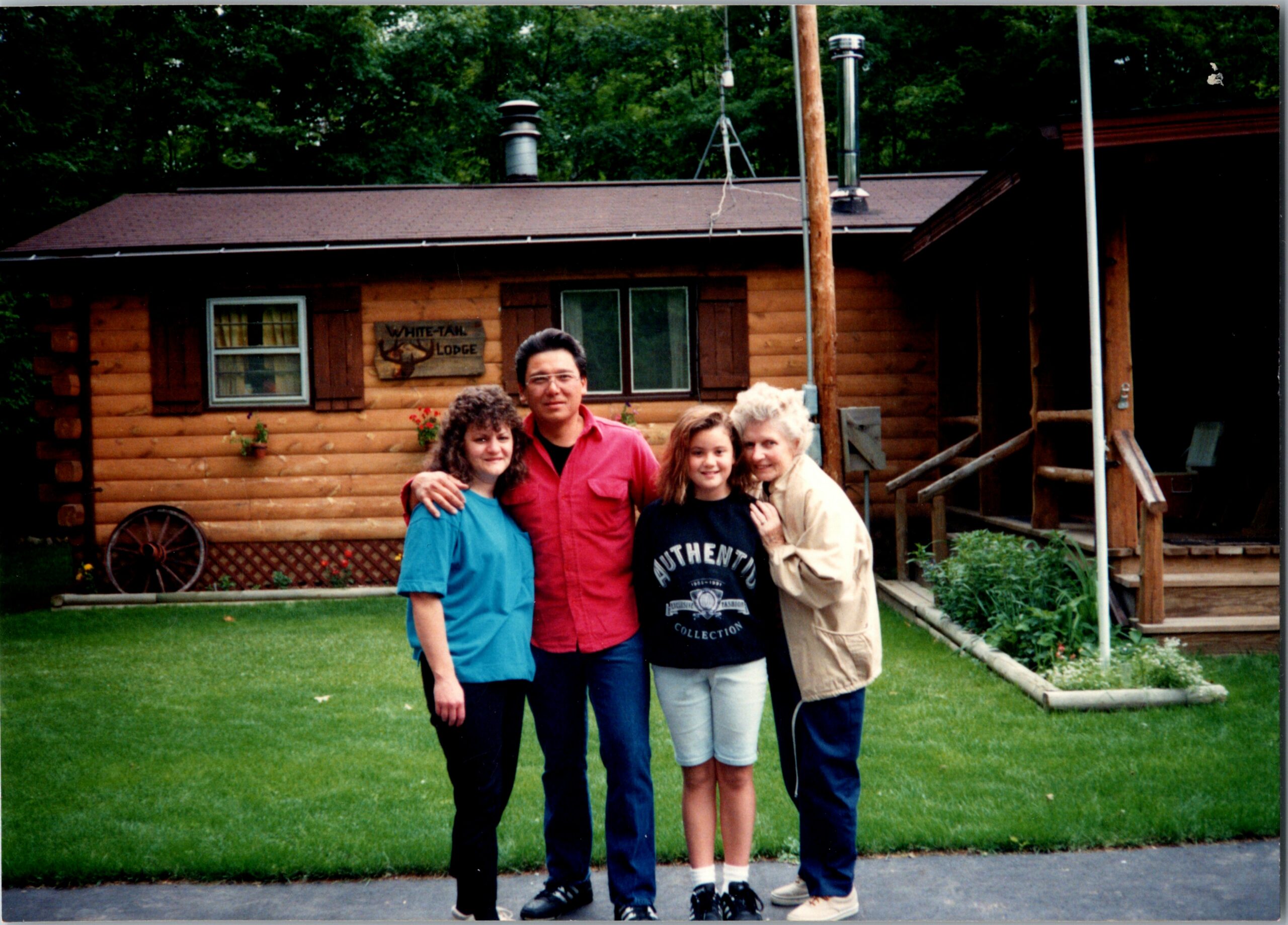 Family of five posing together in front of a log cabin with a grassy yard behind them.
