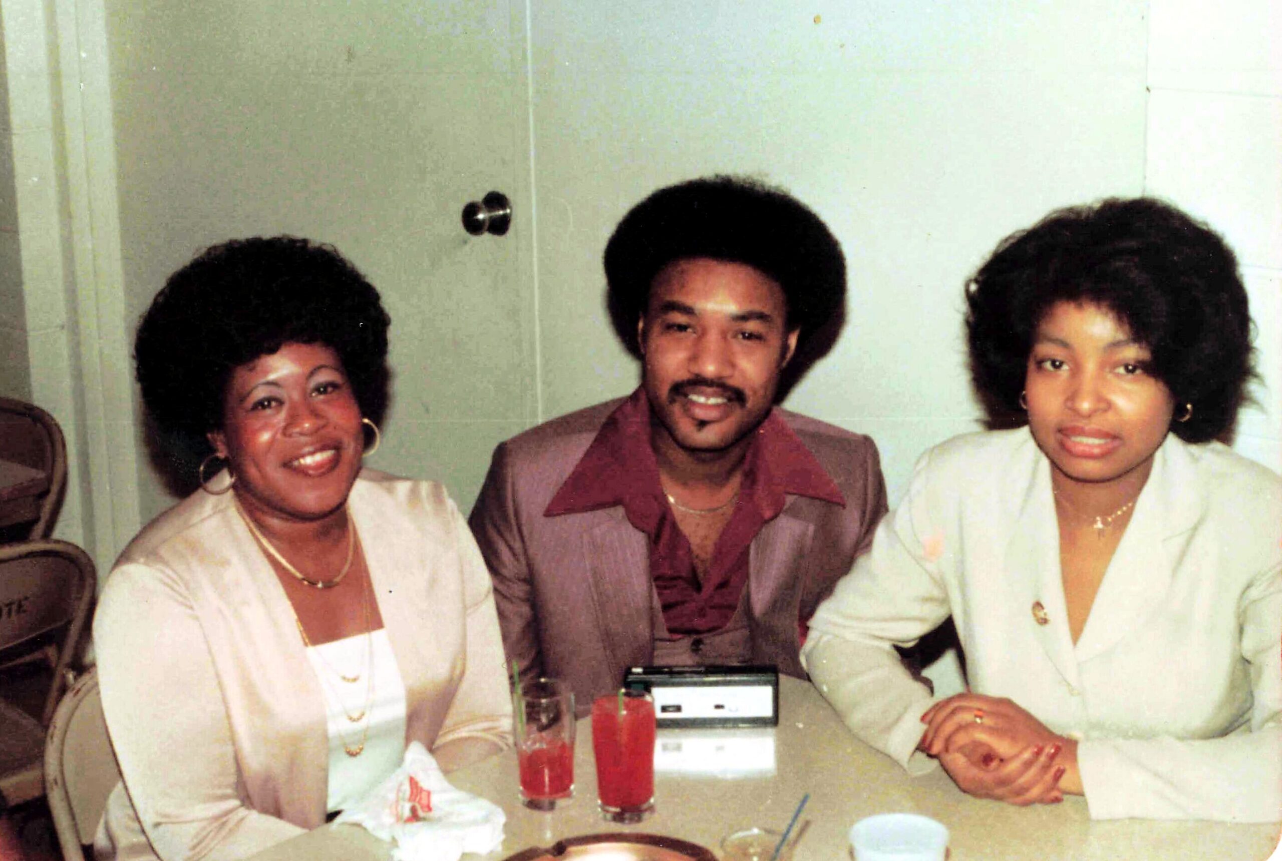 Three friends at a table in a 1970s-style setting, smiling for the camera, wearing light-colored outfits with big hair.
