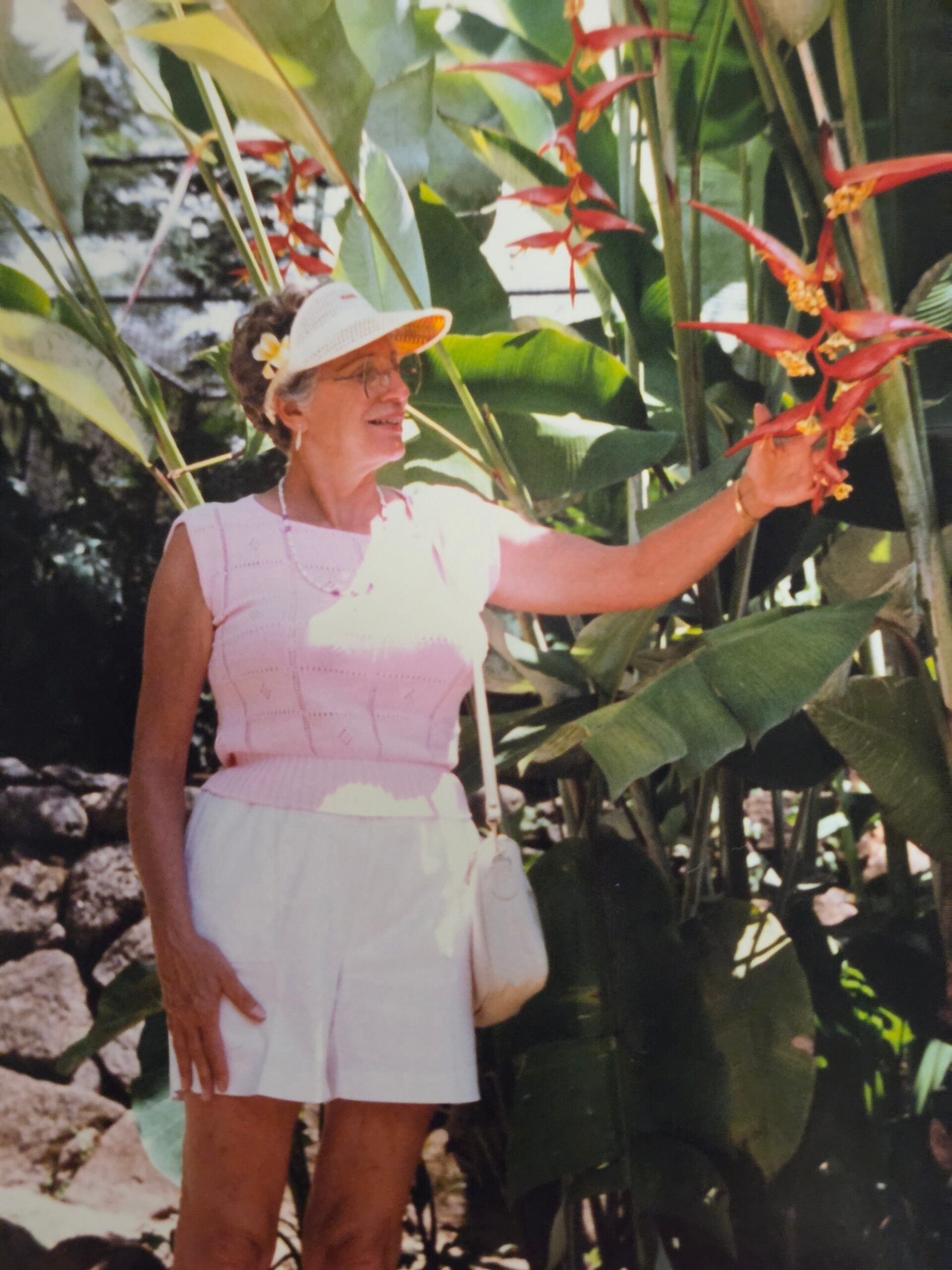 Older woman in a pink sleeveless top and white skirt stands among tropical plants, reaching toward red heliconia flowers.