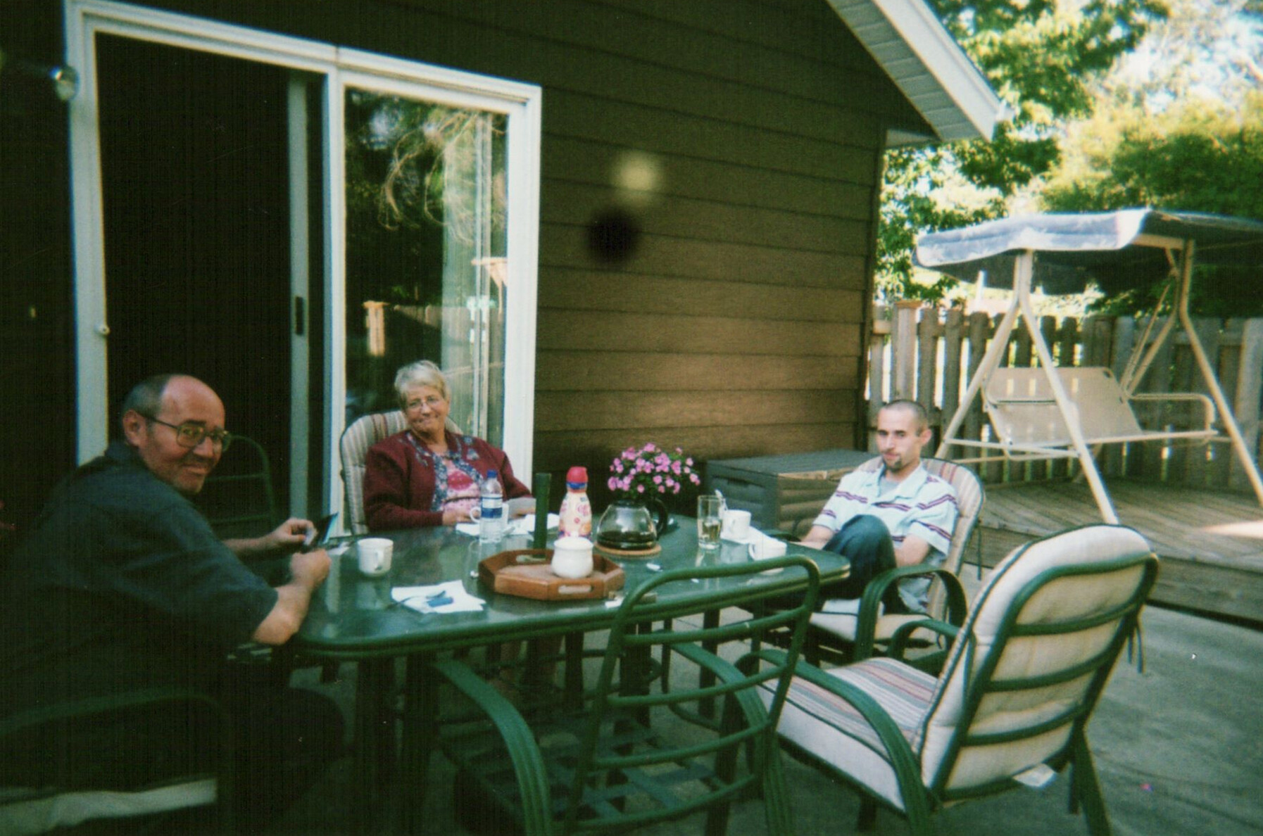 Three adults sit around a glass patio table on a backyard deck, chatting with drinks and mugs visible. A sliding door and brown house wall are in the background, with a wooden fence and a porch swing on the right.