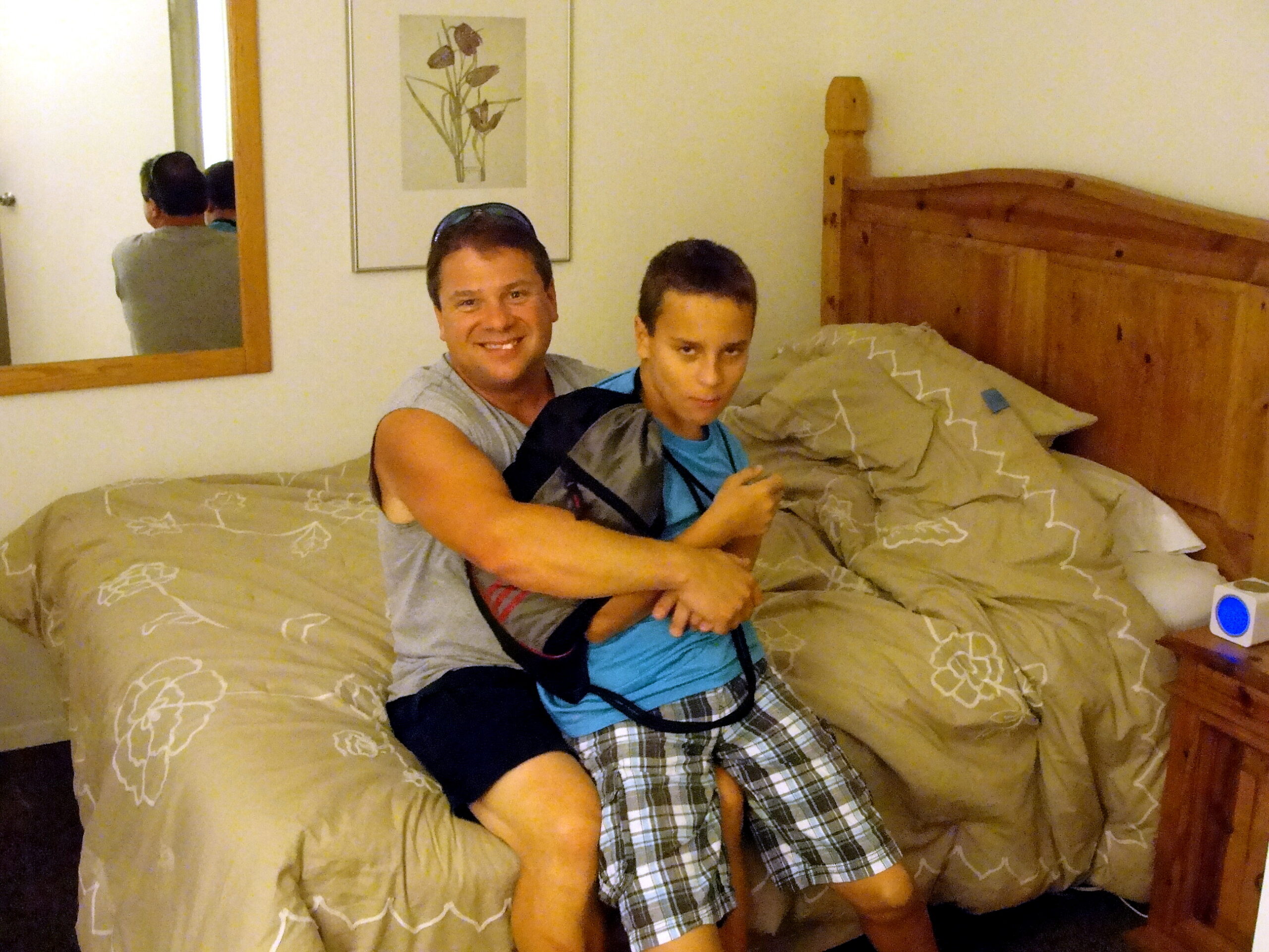 Two men sit on a bed in a beige hotel room, one smiling at the camera while the other looks down; wooden headboard and a framed flower print on the wall behind them.